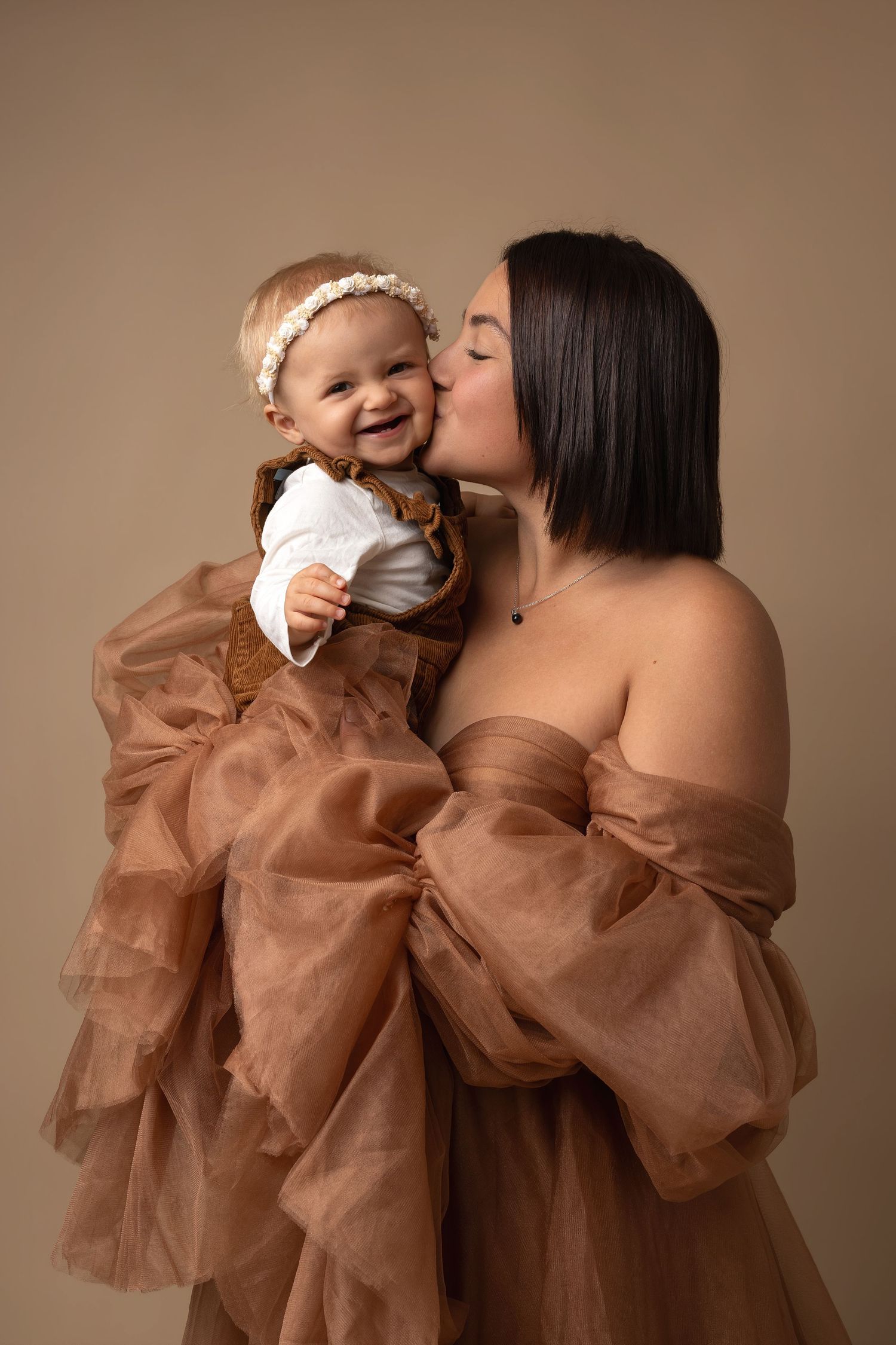 A mother in a brown tulle gown kisses her smiling baby girl wearing a floral headband in a studio portrait.