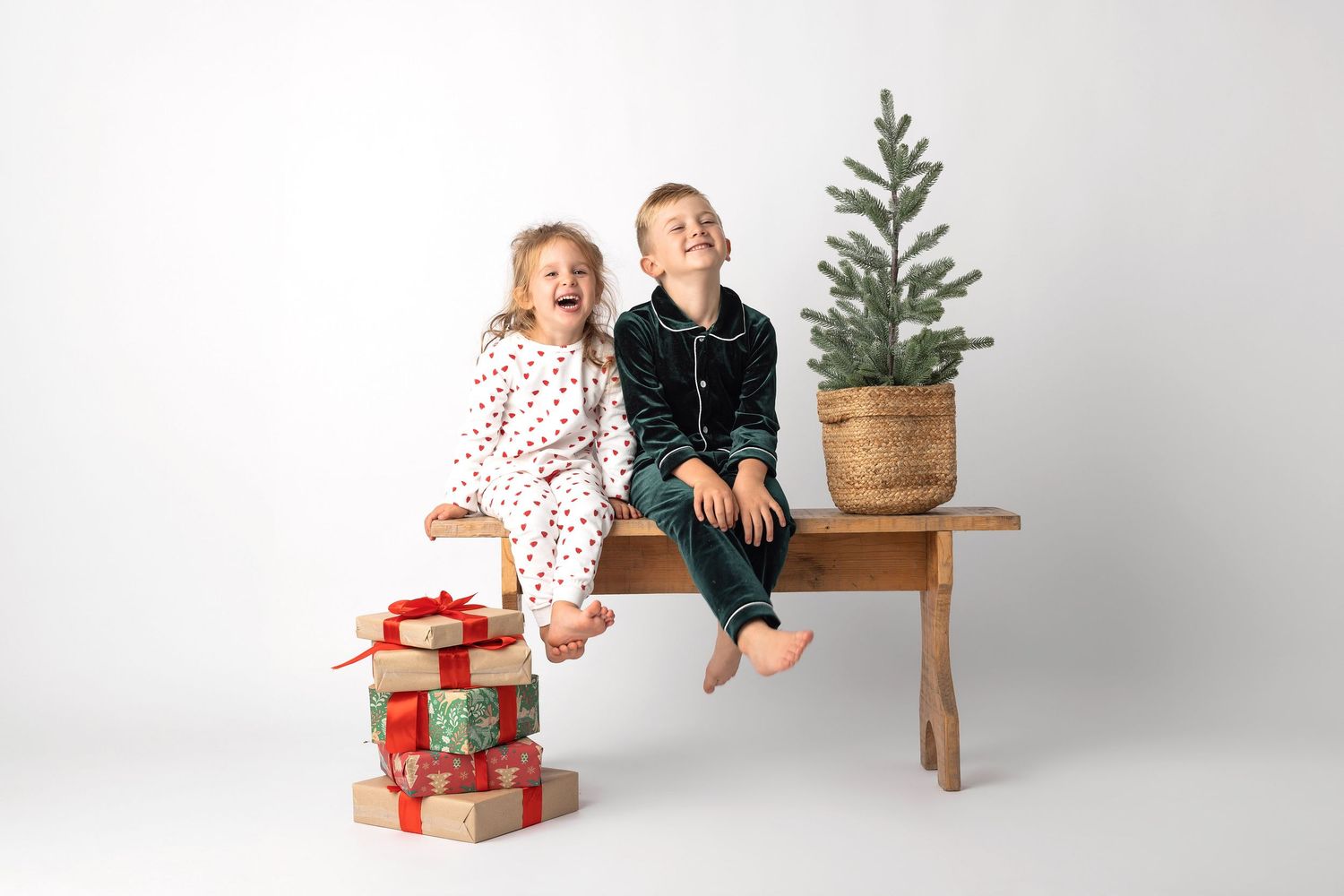 Two joyful children in pajamas sit on a wooden bench with Christmas gifts and a small tree.