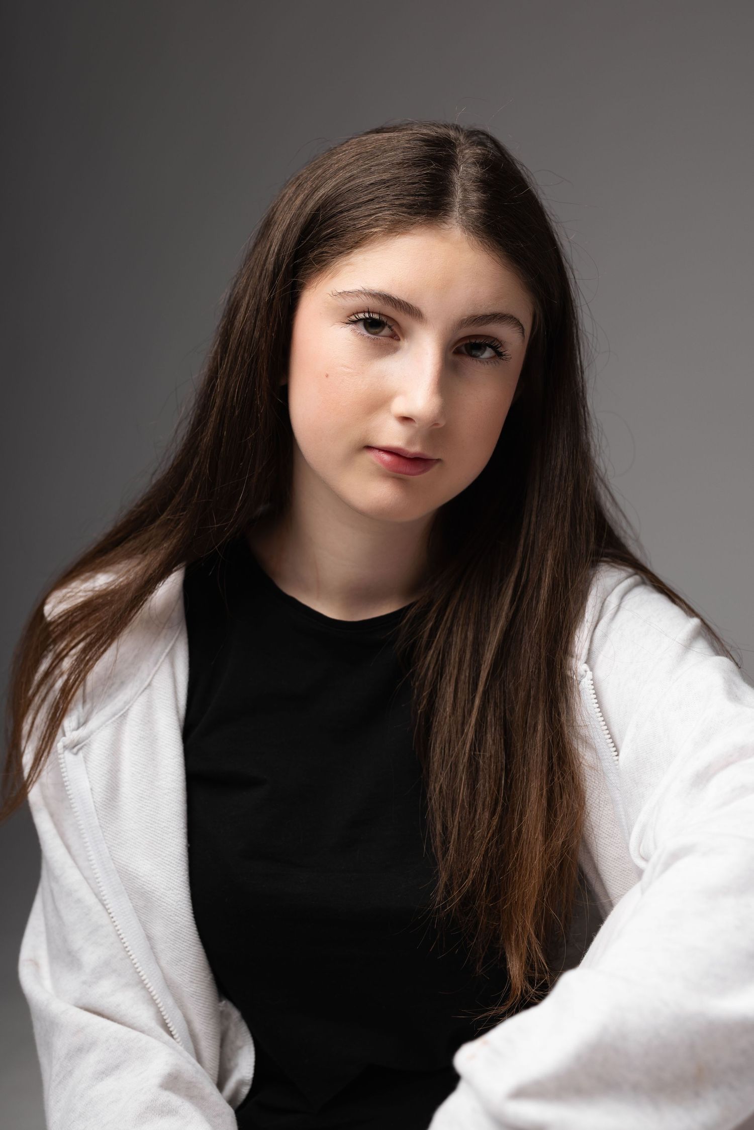 Young woman with long brown hair wearing a black top and white jacket poses against a gray studio background.