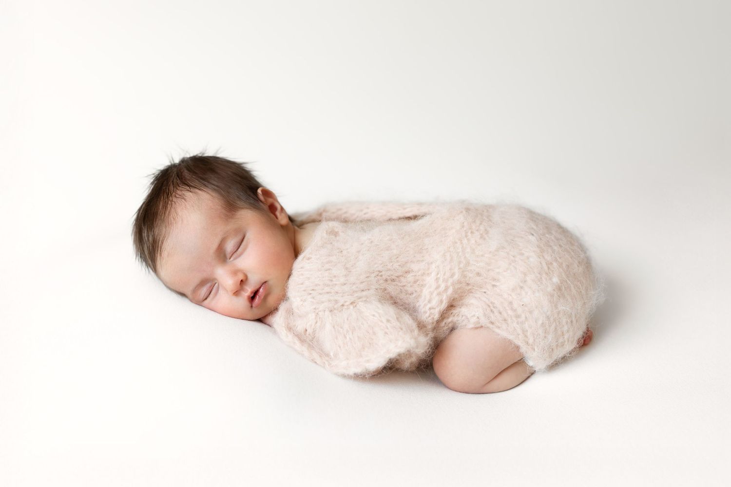 Sleeping newborn baby wrapped in soft pink fuzzy knit romper on white background.