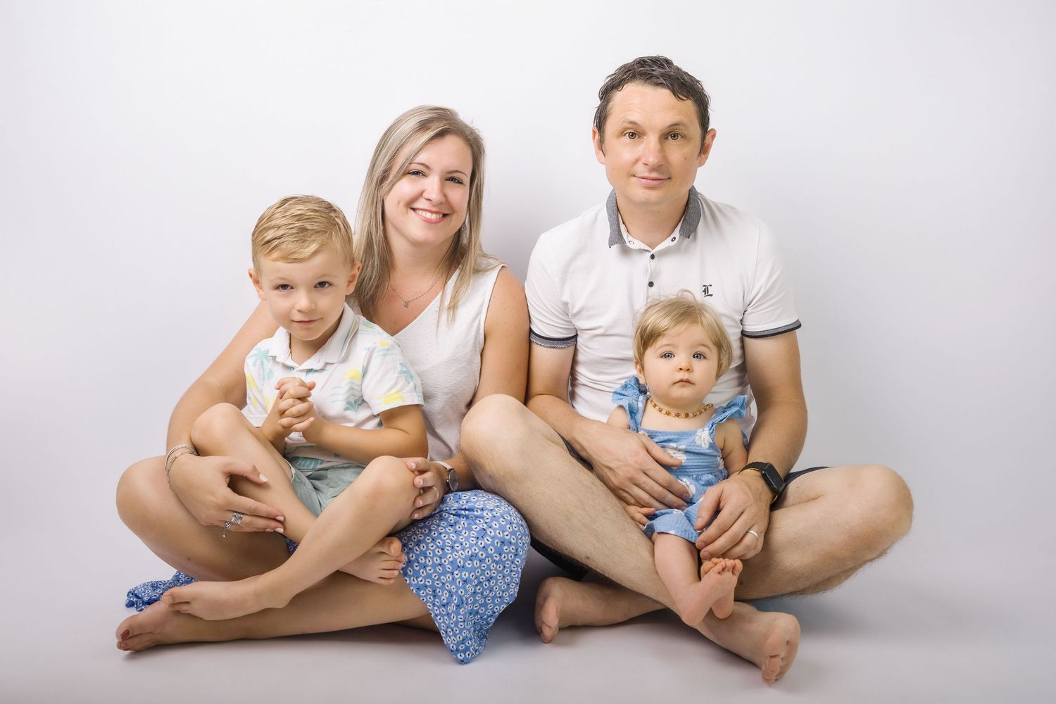 A happy family of four sits together on a white studio floor, parents holding two young children.