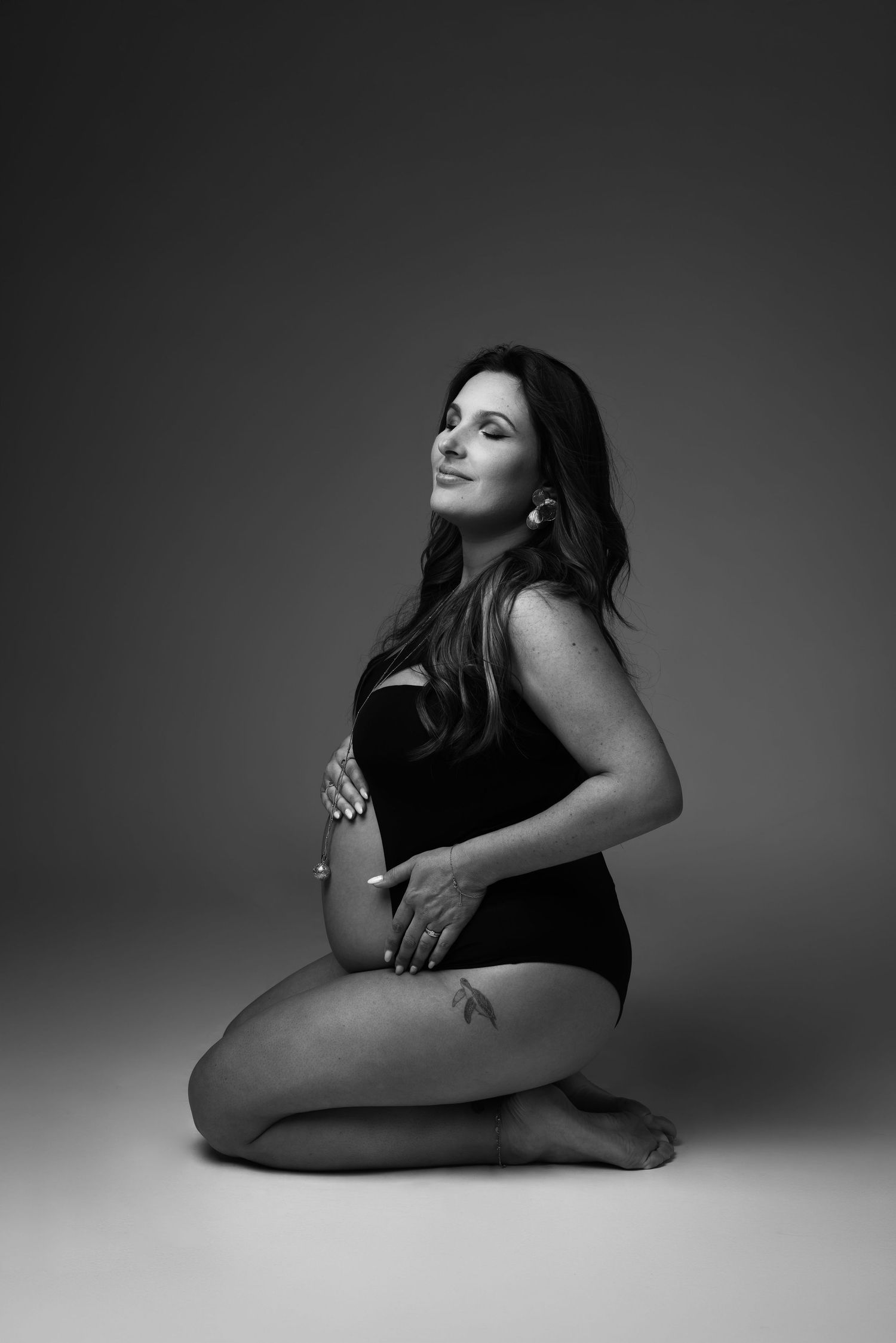 Black and white maternity photo of a pregnant woman in a bodysuit, kneeling gracefully while cradling her baby bump.