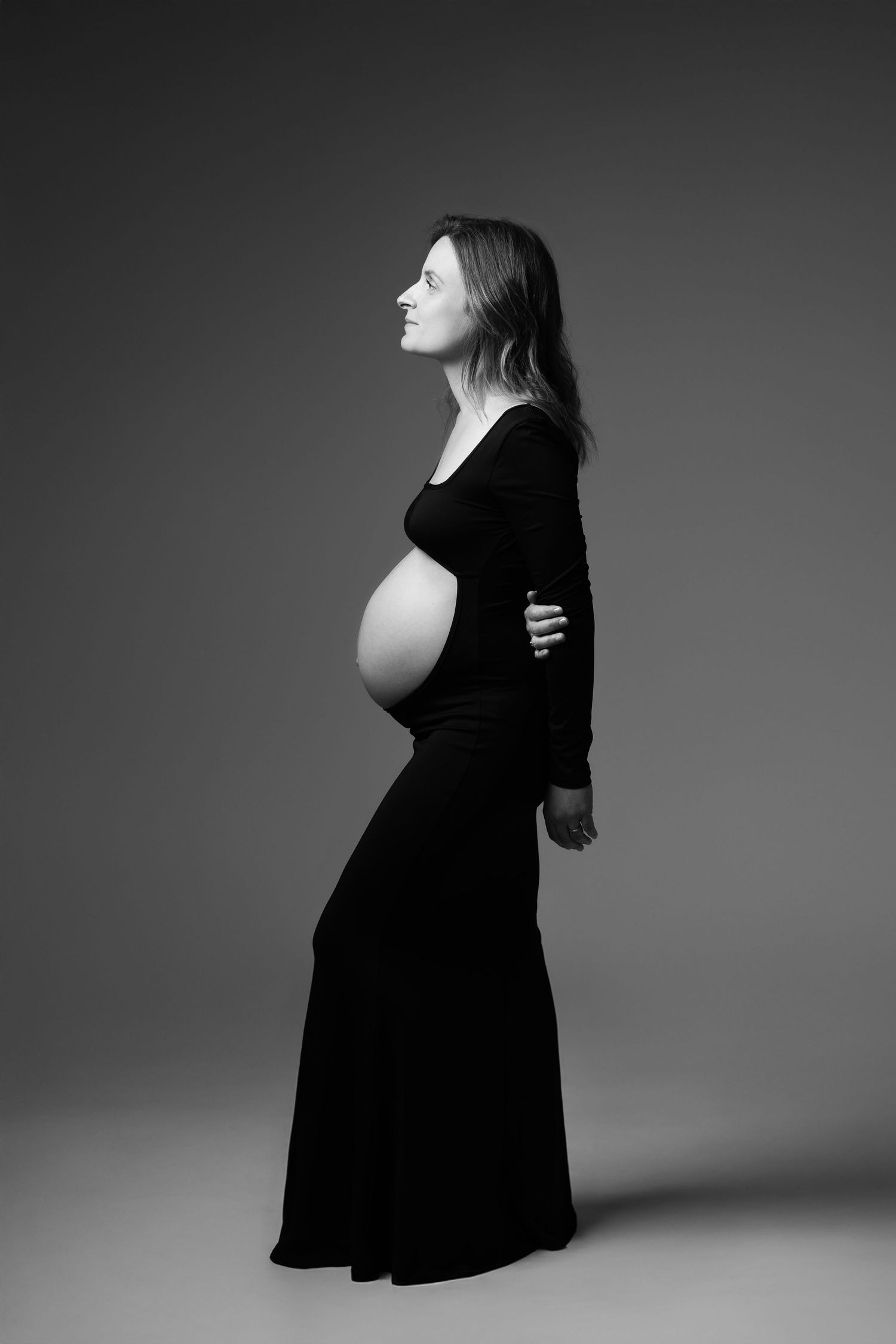 Black and white side profile of pregnant woman in long black dress cradling bump.