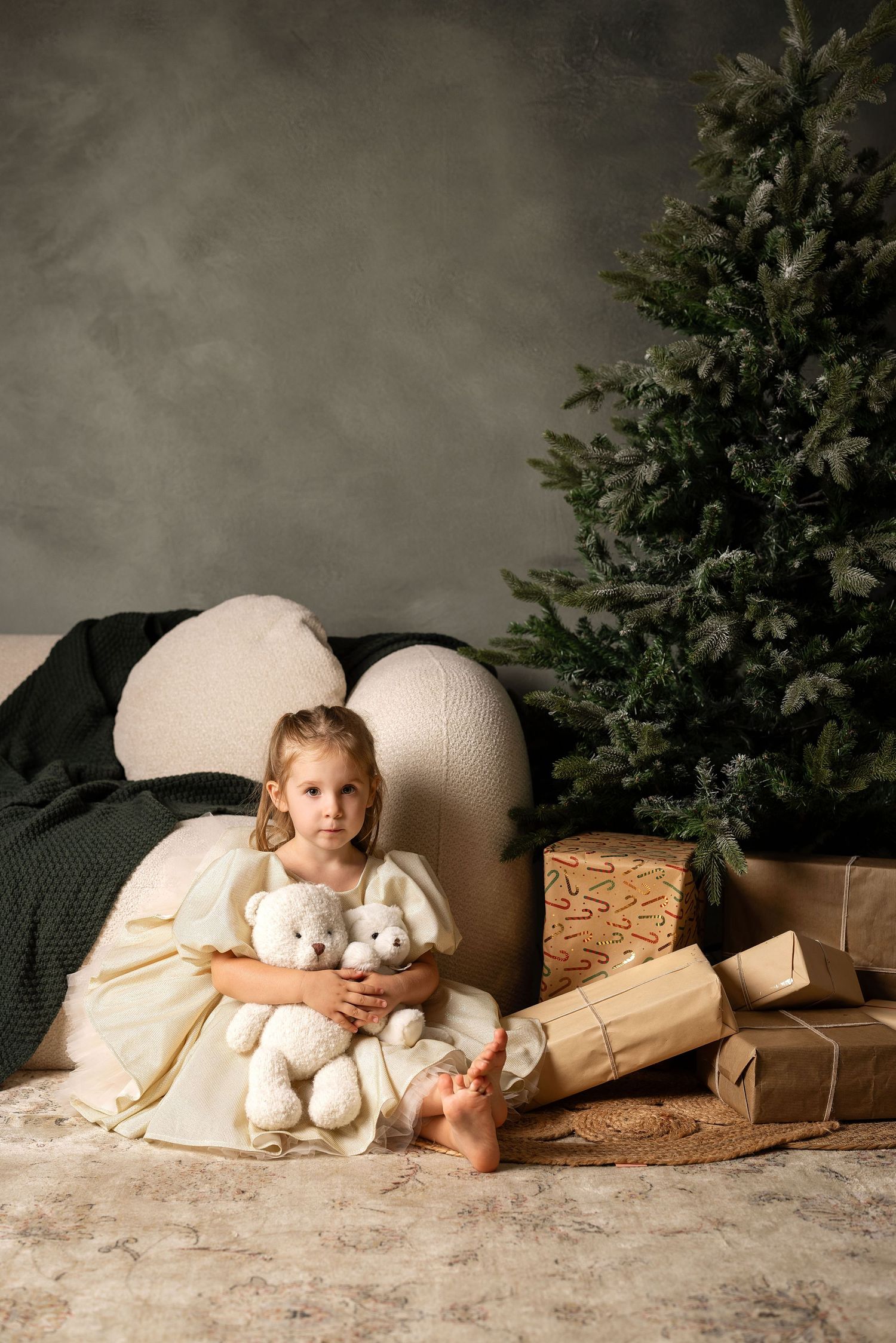 Young girl in white dress hugging stuffed animals beside Christmas tree with wrapped gifts.