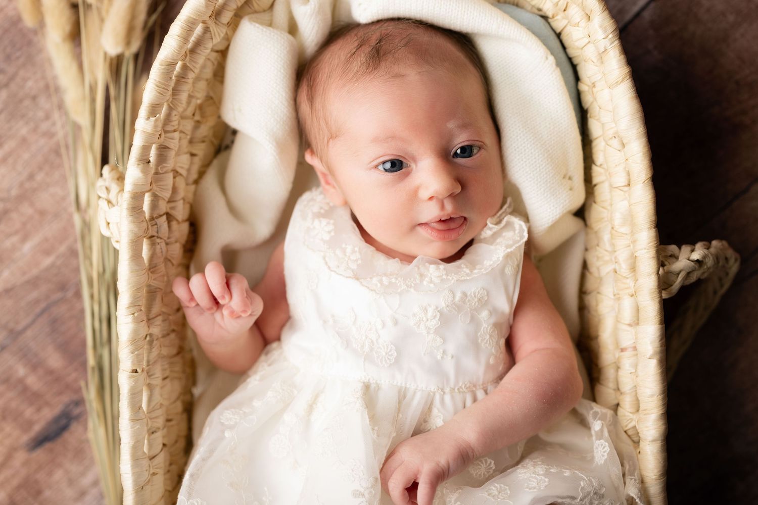 Newborn baby in white lace dress lying in a wicker basket with soft white lining, looking up with blue eyes.