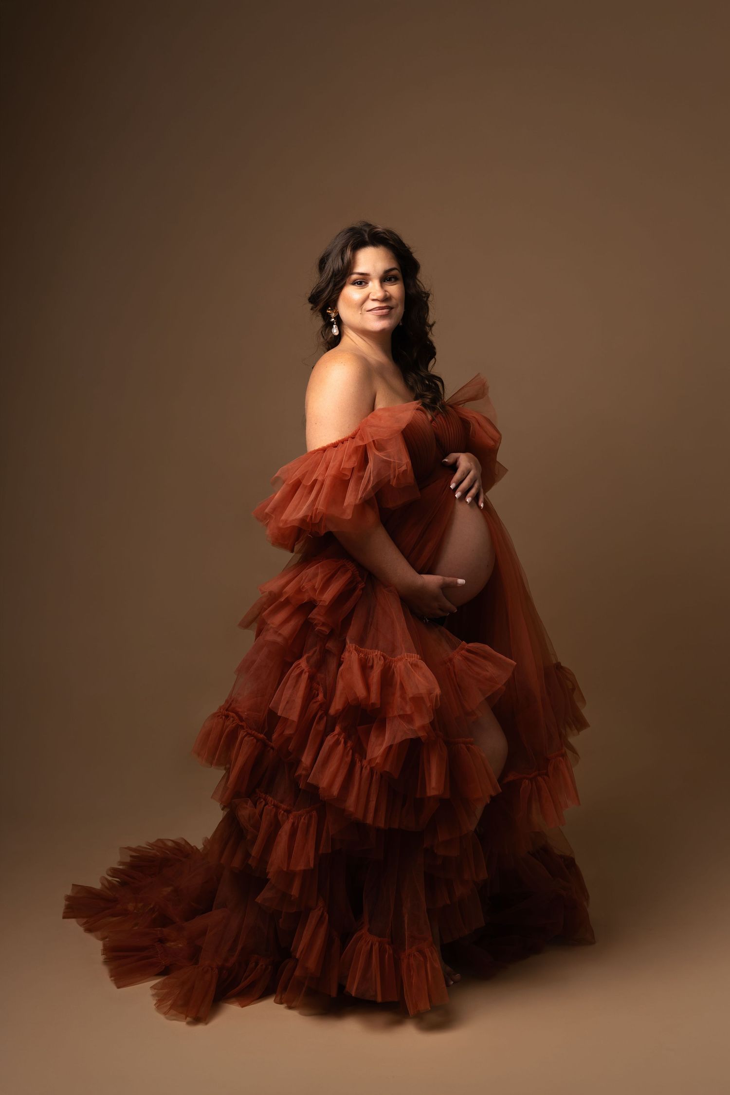 Pregnant woman in a rust-colored ruffled tulle gown cradling her baby bump during a maternity photo session.