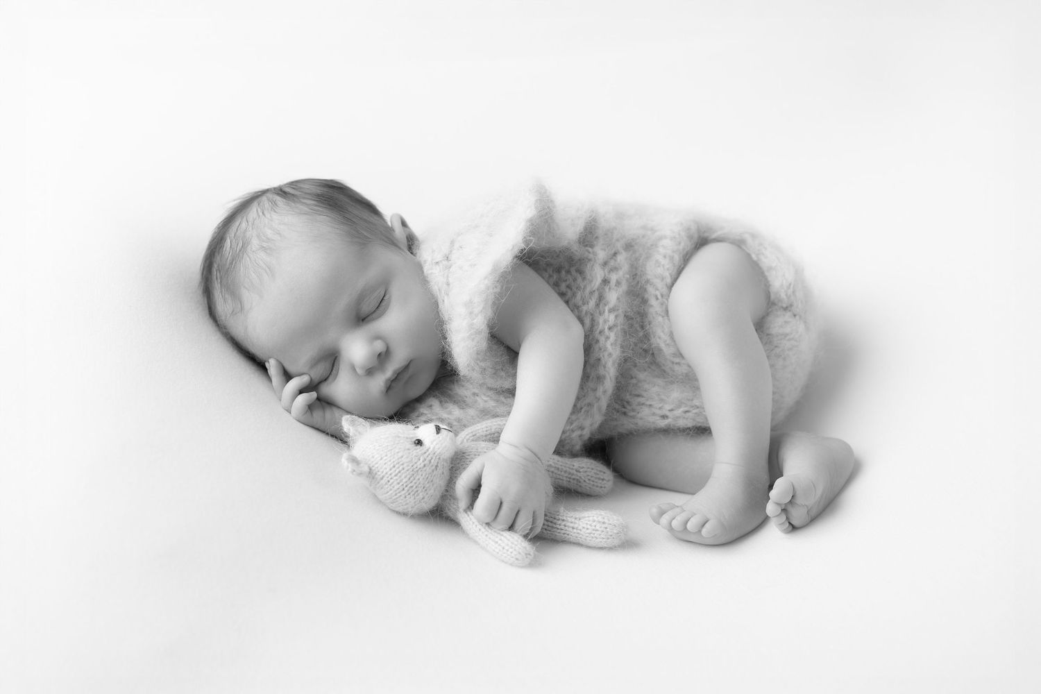 Black and white photo of sleeping newborn baby in knit romper cuddling a small stuffed teddy bear.