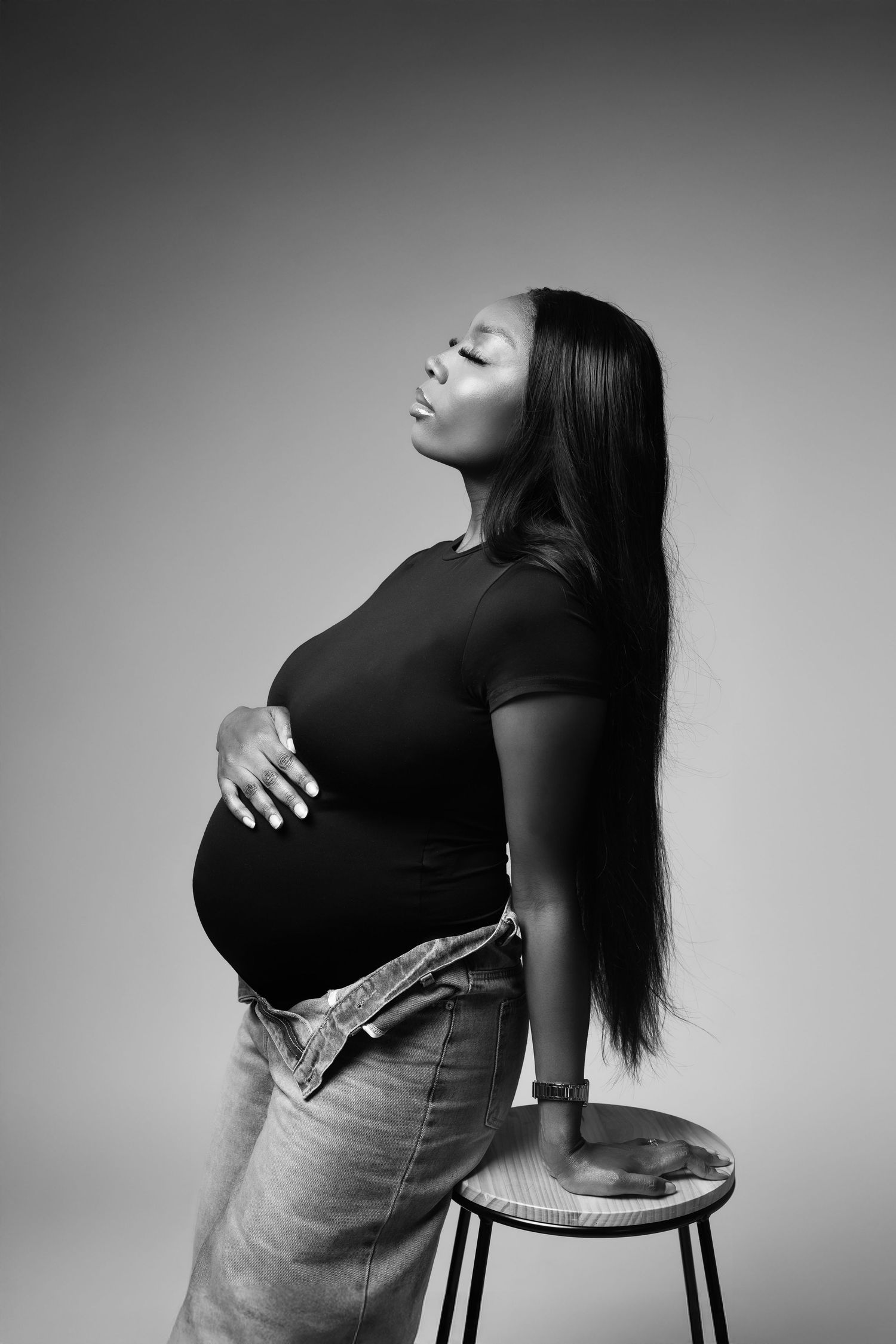 Black and white portrait of pregnant woman leaning on stool in studio.