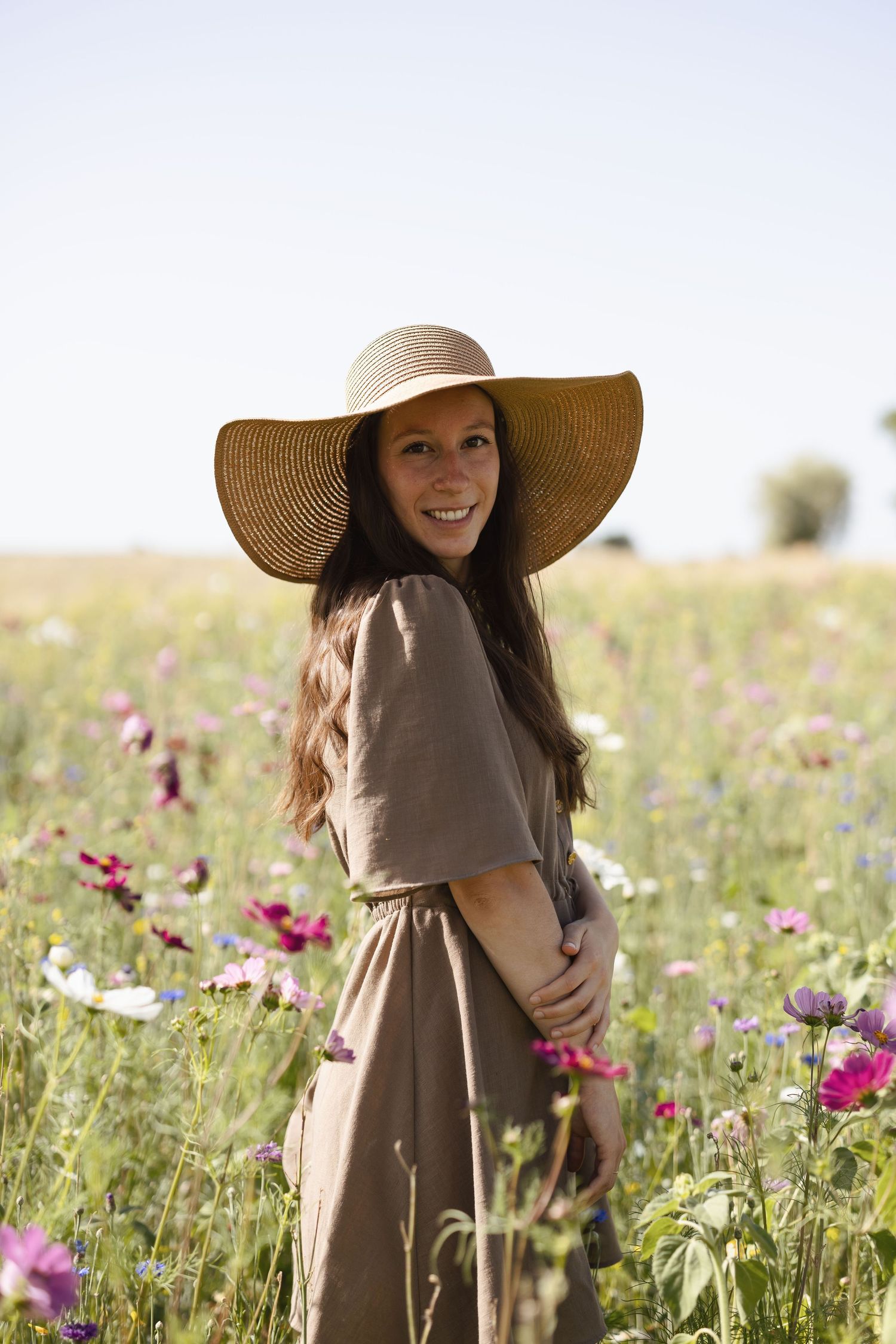 Smiling woman in wide-brim straw hat and brown dress standing in a colorful wildflower field on a sunny day.