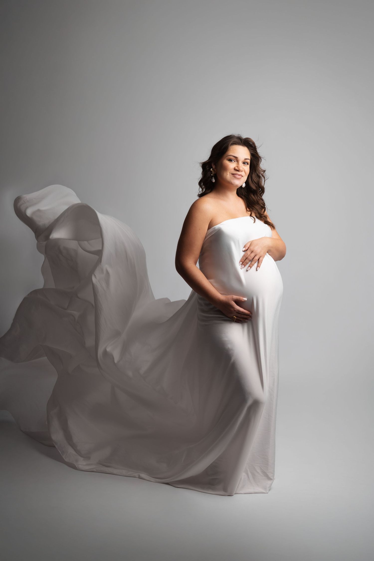 Pregnant woman in flowing white fabric gown cradling baby bump, studio maternity photo on gray background.