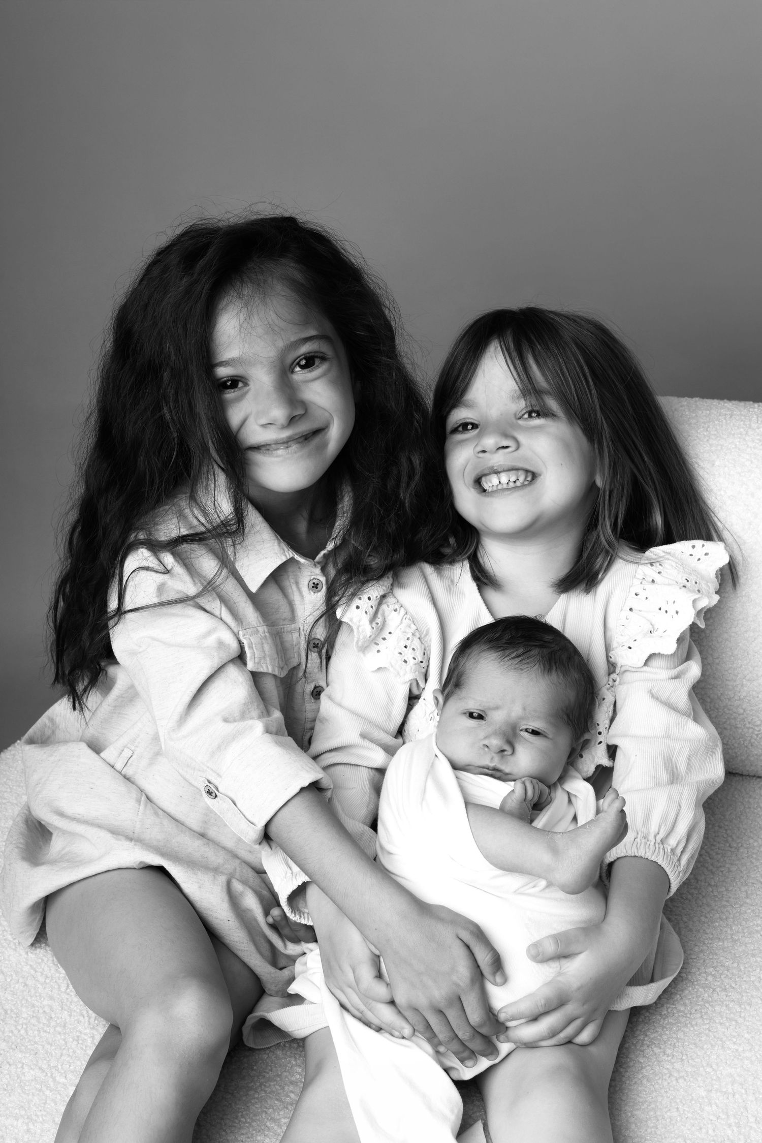 Black and white portrait of two smiling girls holding a newborn baby, seated together in a chair.