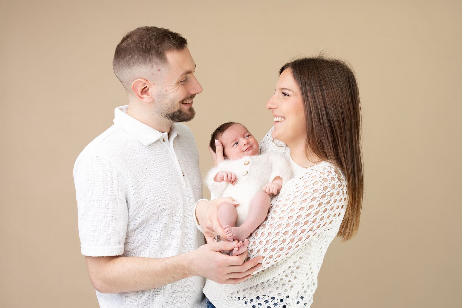 Smiling parents in white outfits holding their newborn baby together against beige background.