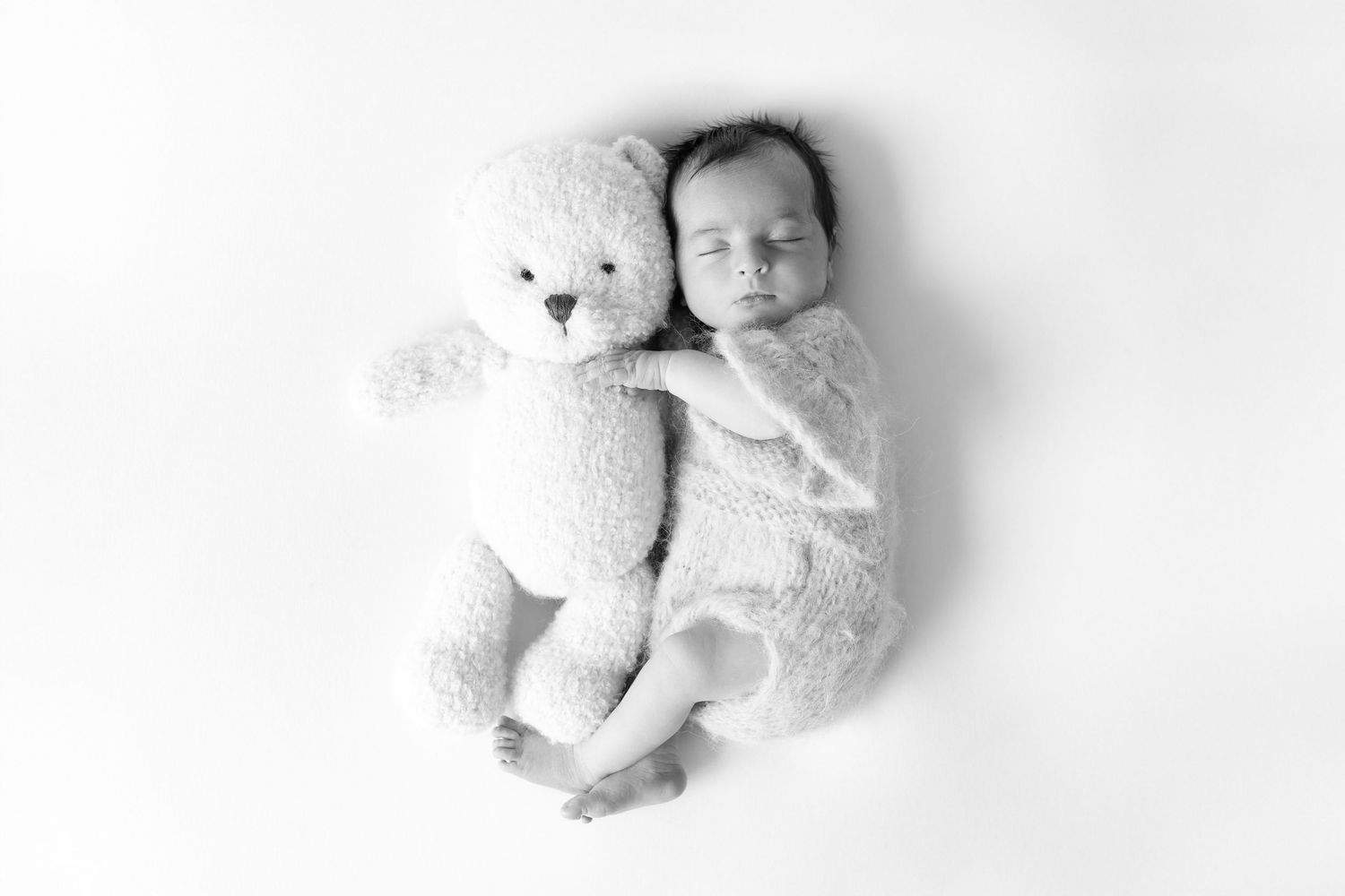 Sleeping newborn baby cuddled next to a large teddy bear in black and white photo.