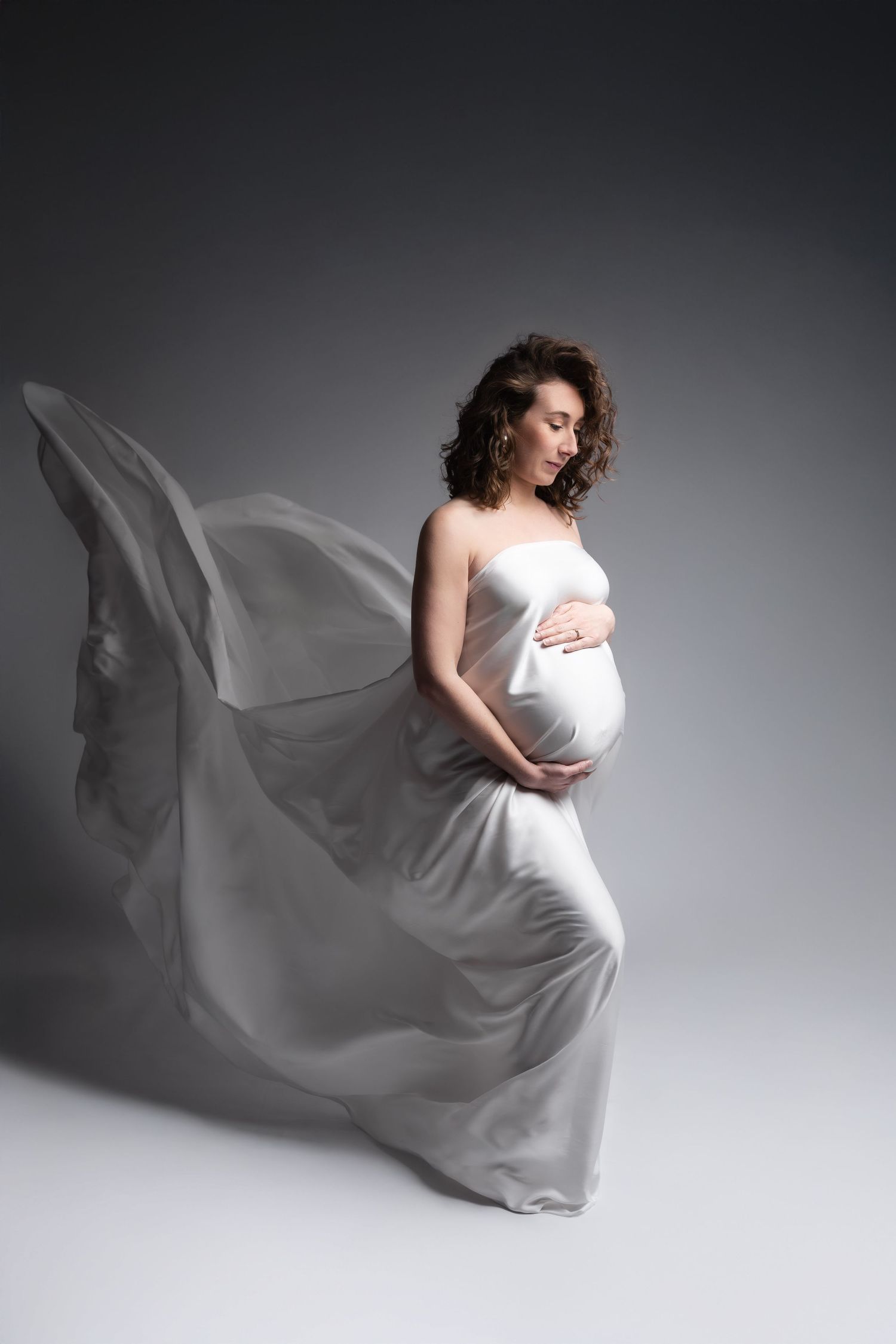 Pregnant woman in flowing white silk fabric dress cradles her bump, eyes closed, on a grey studio background.