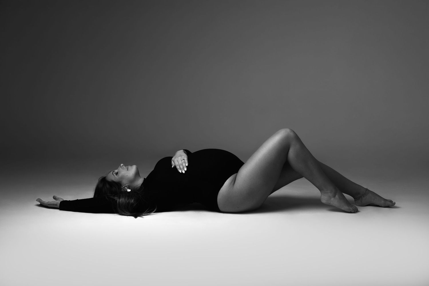 Black and white photo of a pregnant woman in a bodysuit lying gracefully on the floor in a studio setting.