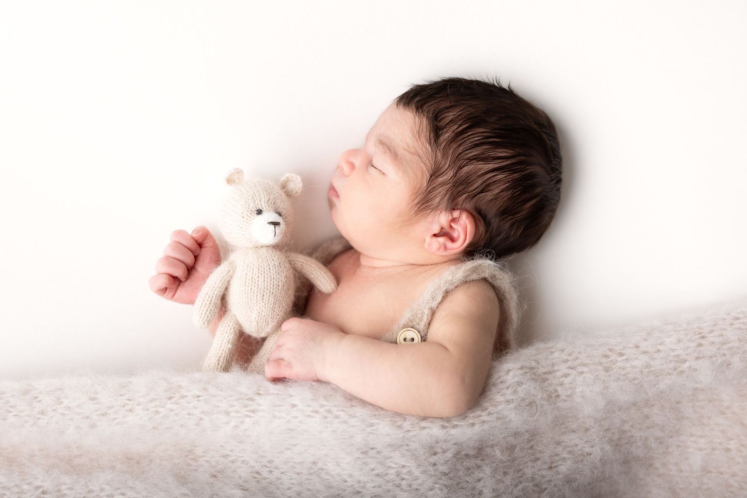 Peaceful newborn baby sleeping while holding a small knitted teddy bear on a soft white blanket. photographe naissance chavanoz lyon