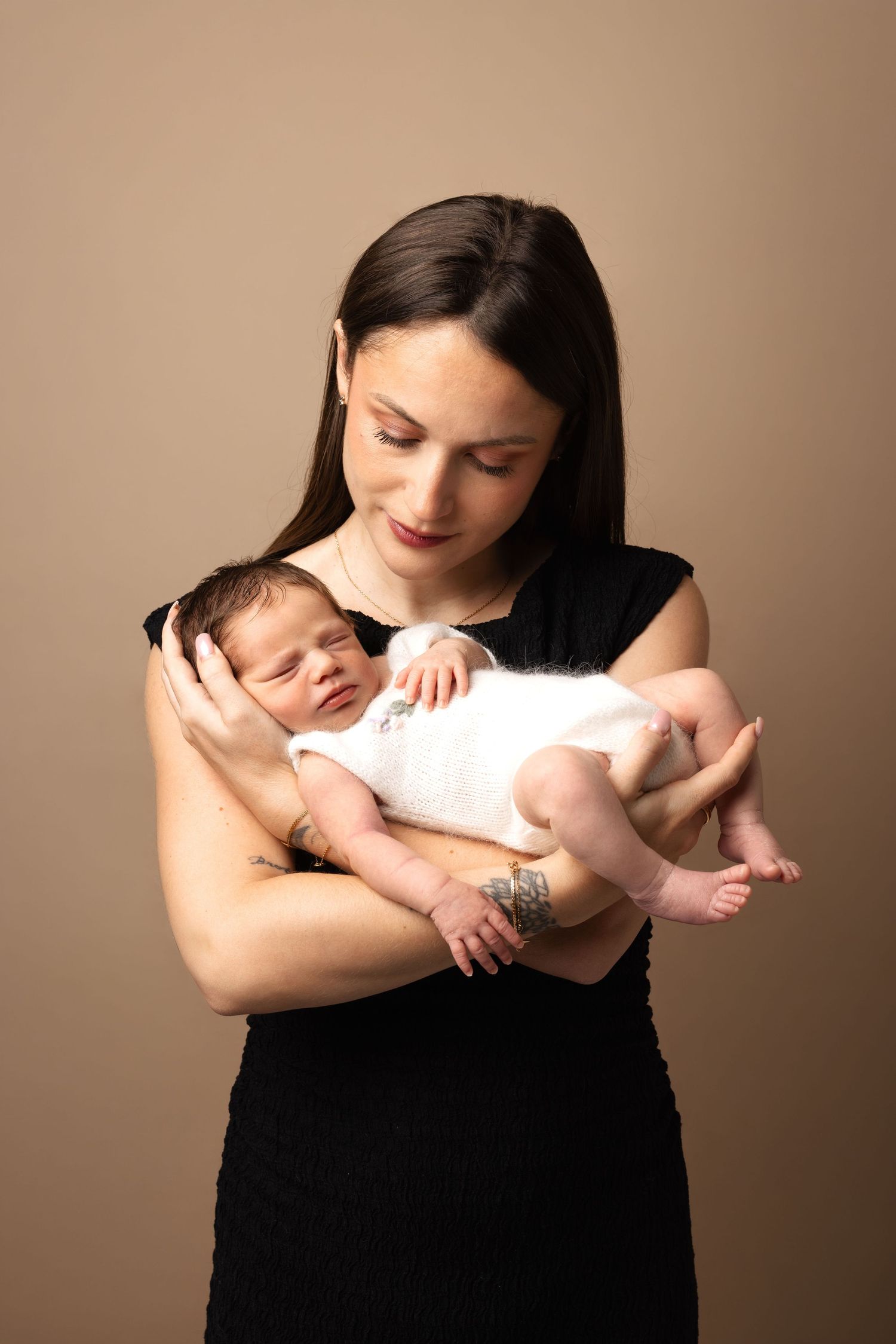 A young mother in a black dress tenderly holds her sleeping newborn baby dressed in white.