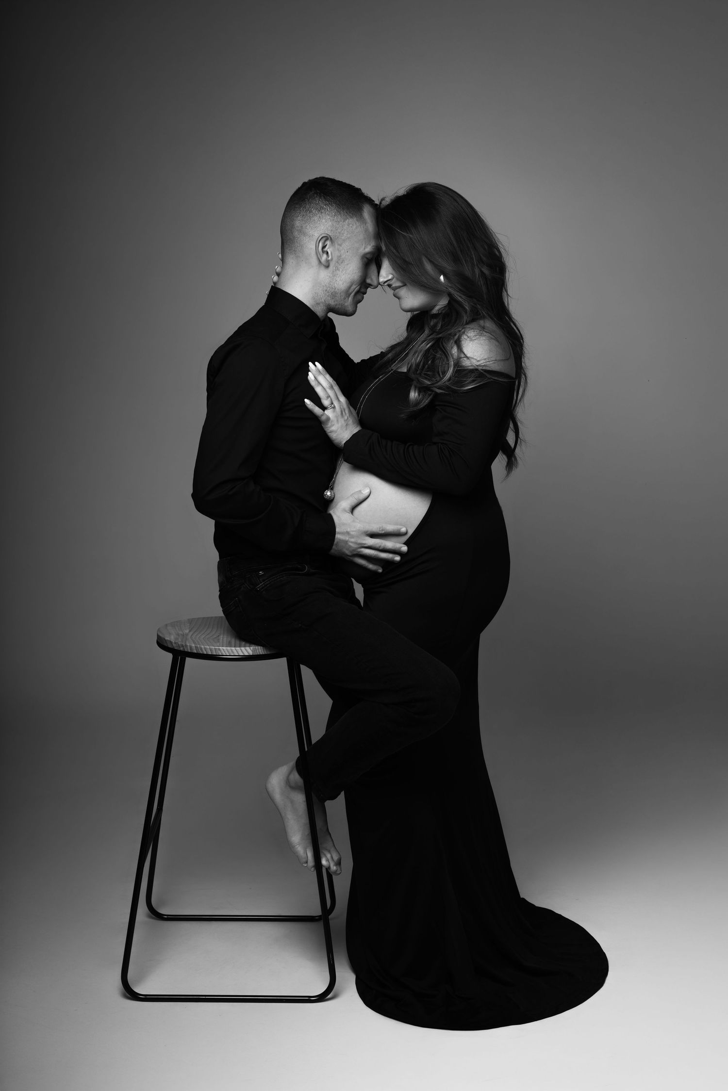 Black and white maternity photo of couple in black outfits, man seated on stool embracing pregnant woman, foreheads touching.