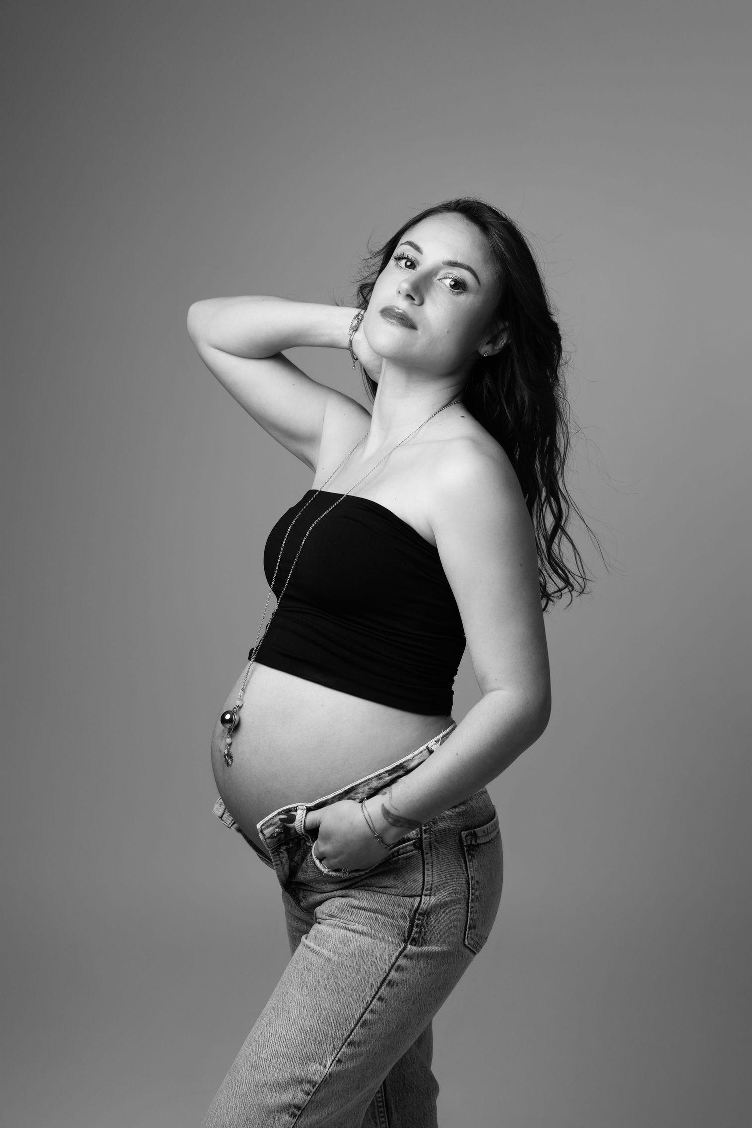 Black and white portrait of a pregnant woman in a crop top and jeans posing gracefully in a studio.