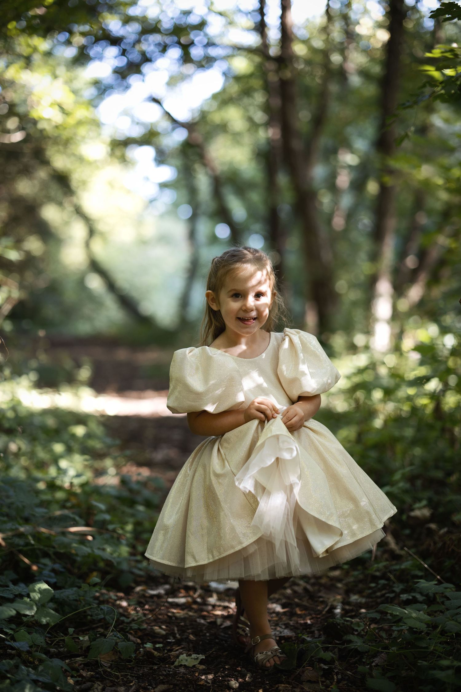 A smiling little girl in a cream puff-sleeve dress runs along a sunlit forest path surrounded by lush green trees.