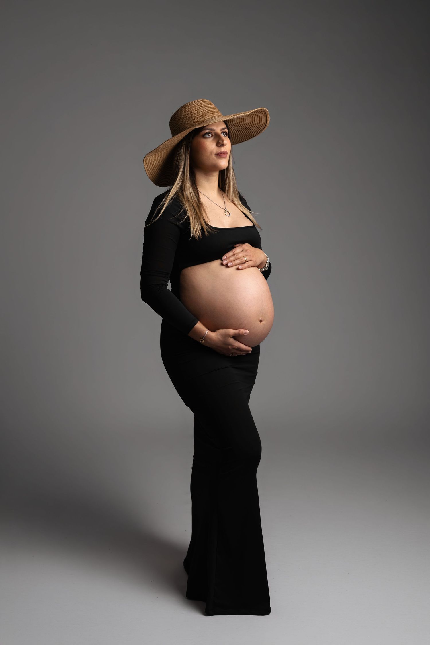 Pregnant woman in black outfit and wide-brim hat cradling baby bump in studio portrait.