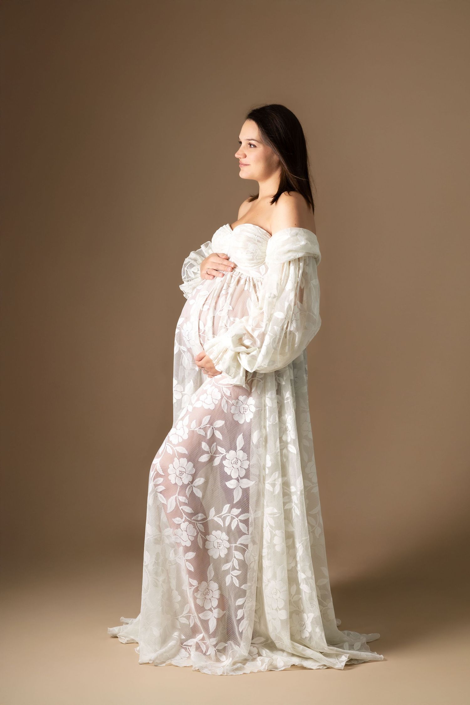 Pregnant woman in an elegant sheer white floral lace gown cradling her baby bump in a studio portrait.