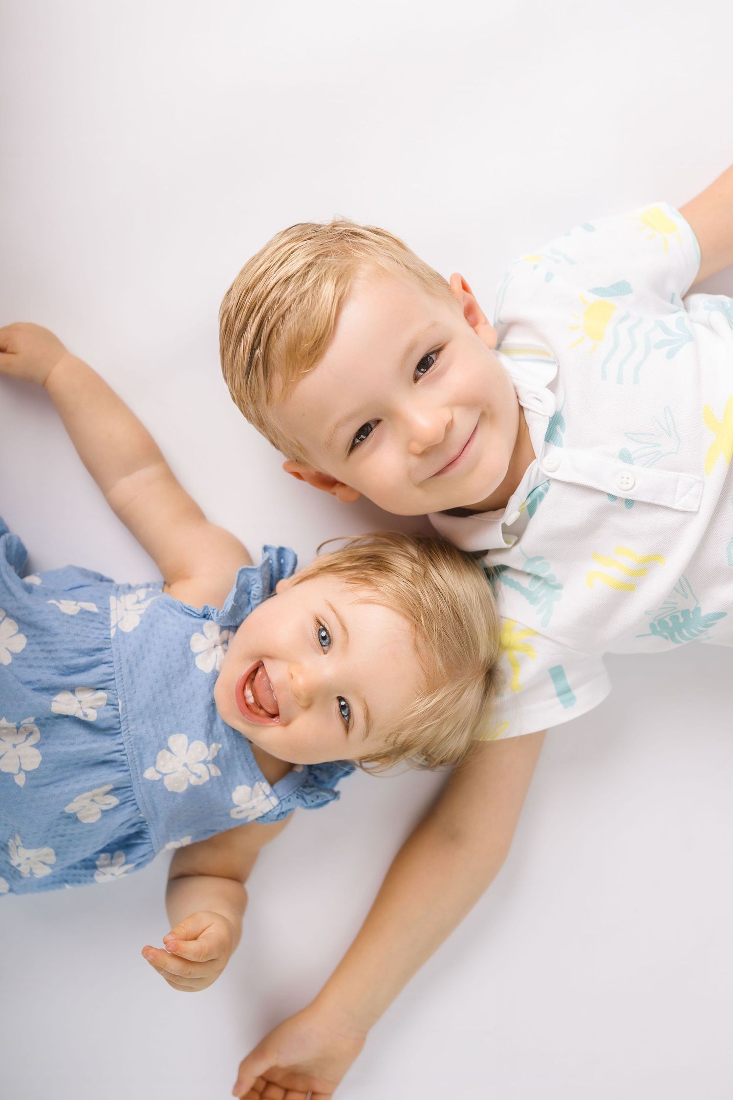 Two happy blonde children lay together on a white background, smiling up at the camera.