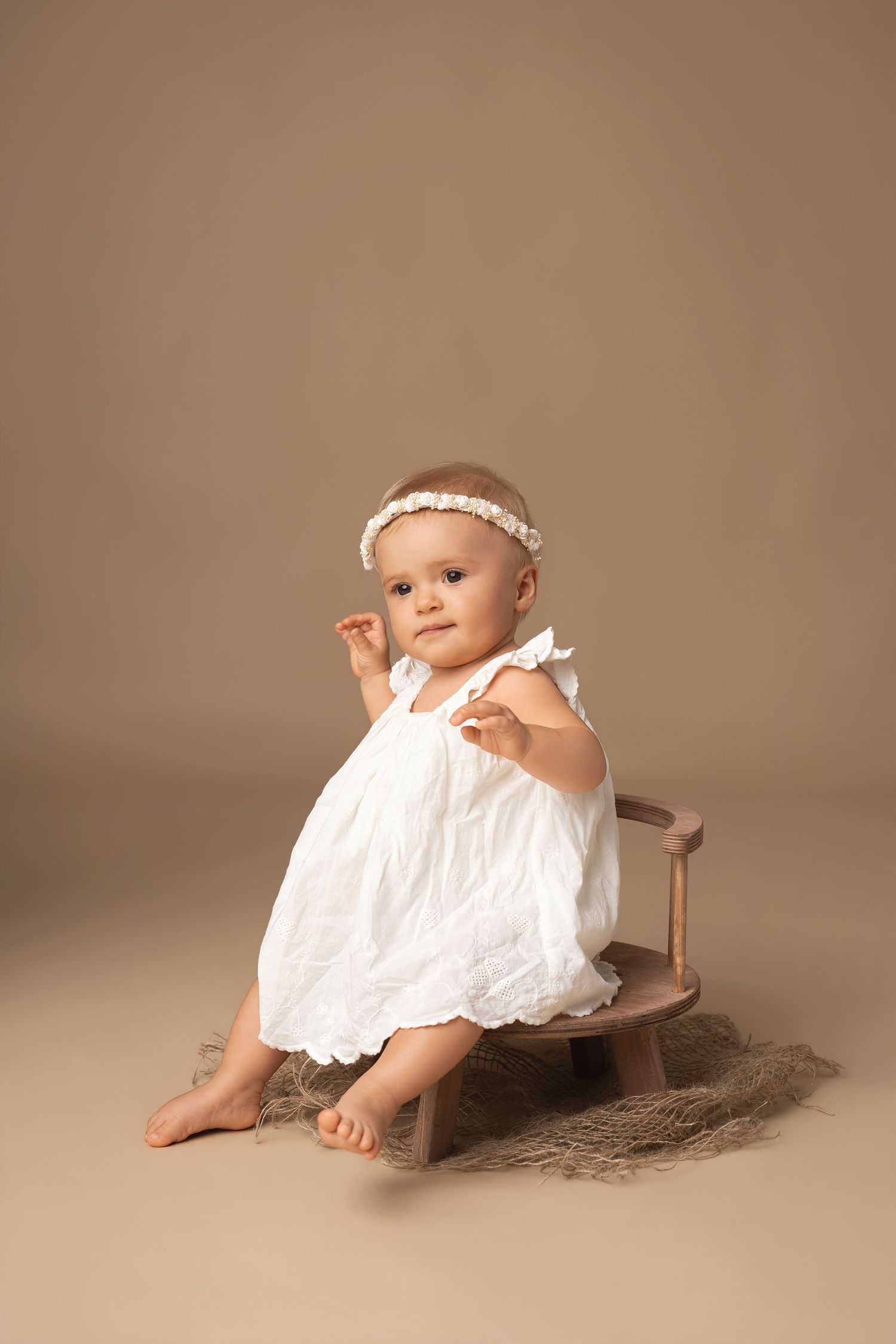 A baby girl in a white dress and headband sits on a small wooden bench against a beige background.