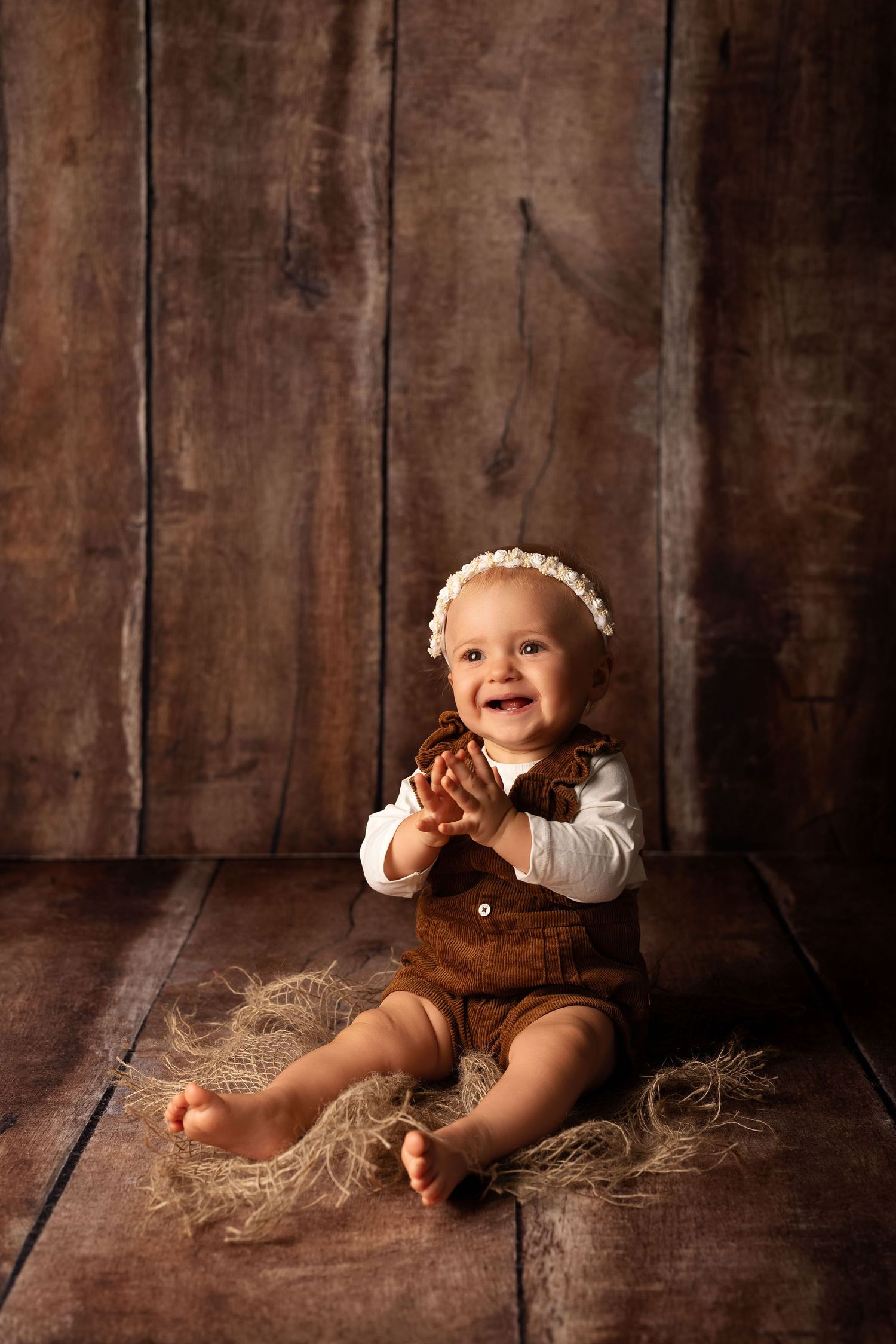 Smiling baby in brown overalls and lace headband clapping hands, sitting on rustic wood floor with hay.