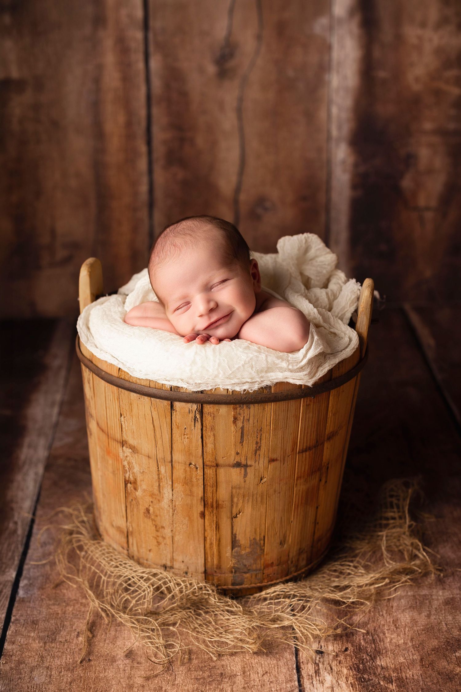 Sleeping newborn baby resting in a rustic wooden bucket with soft white fabric on a wooden floor.