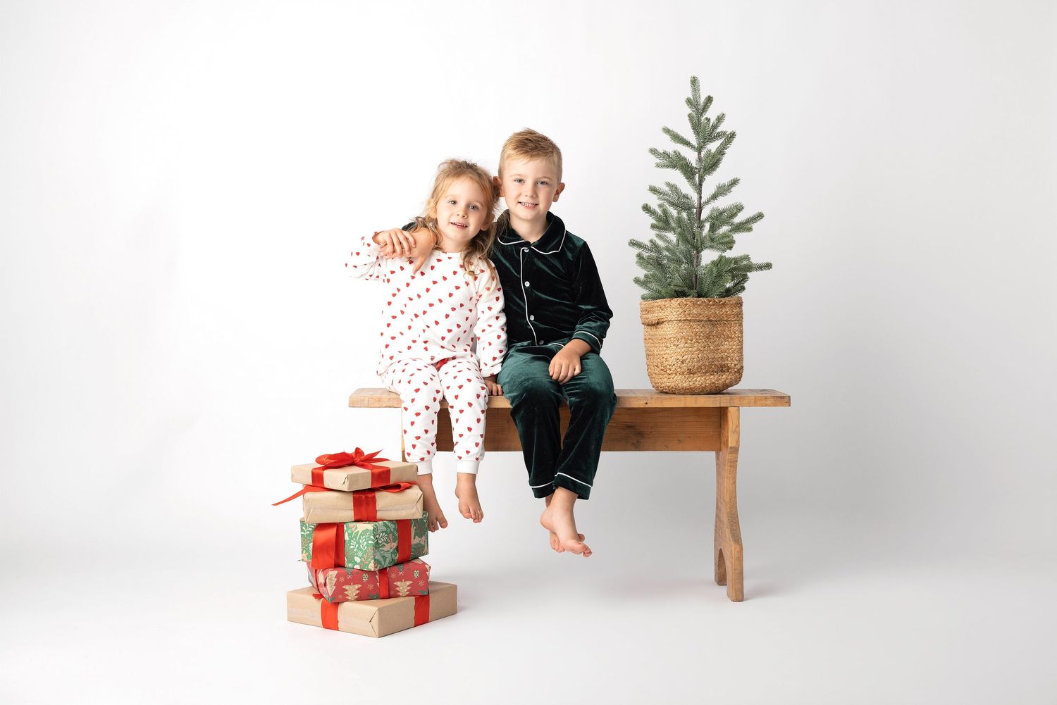 Two children in Christmas pajamas sitting on a wooden bench with wrapped gifts and a small pine tree.