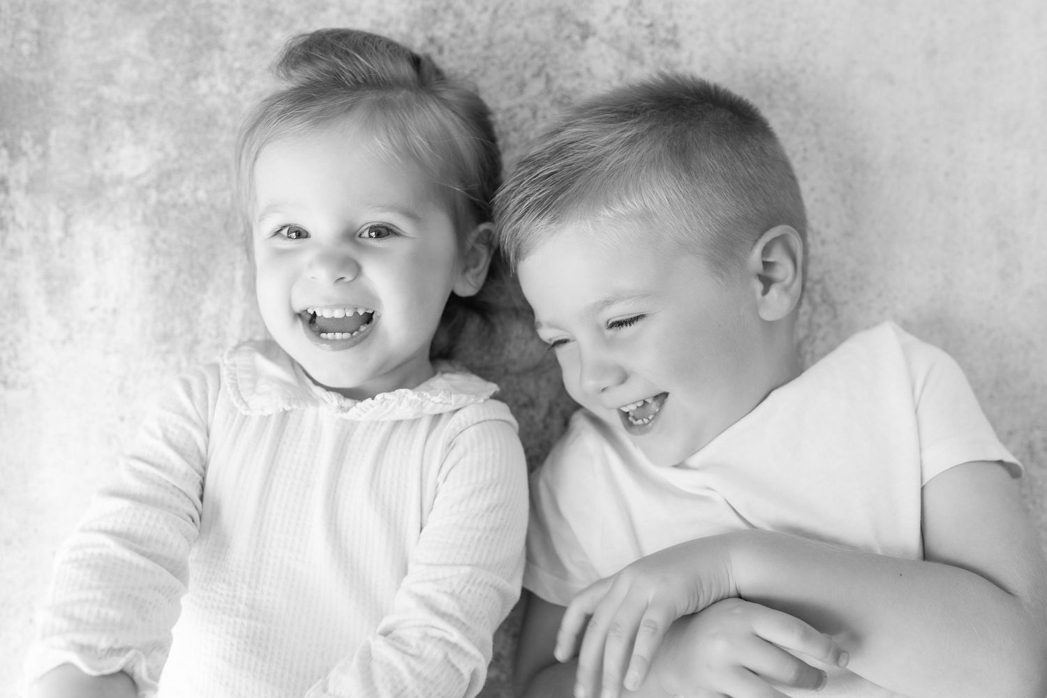 Two happy siblings, a toddler girl and older boy, laugh together in a black and white portrait.