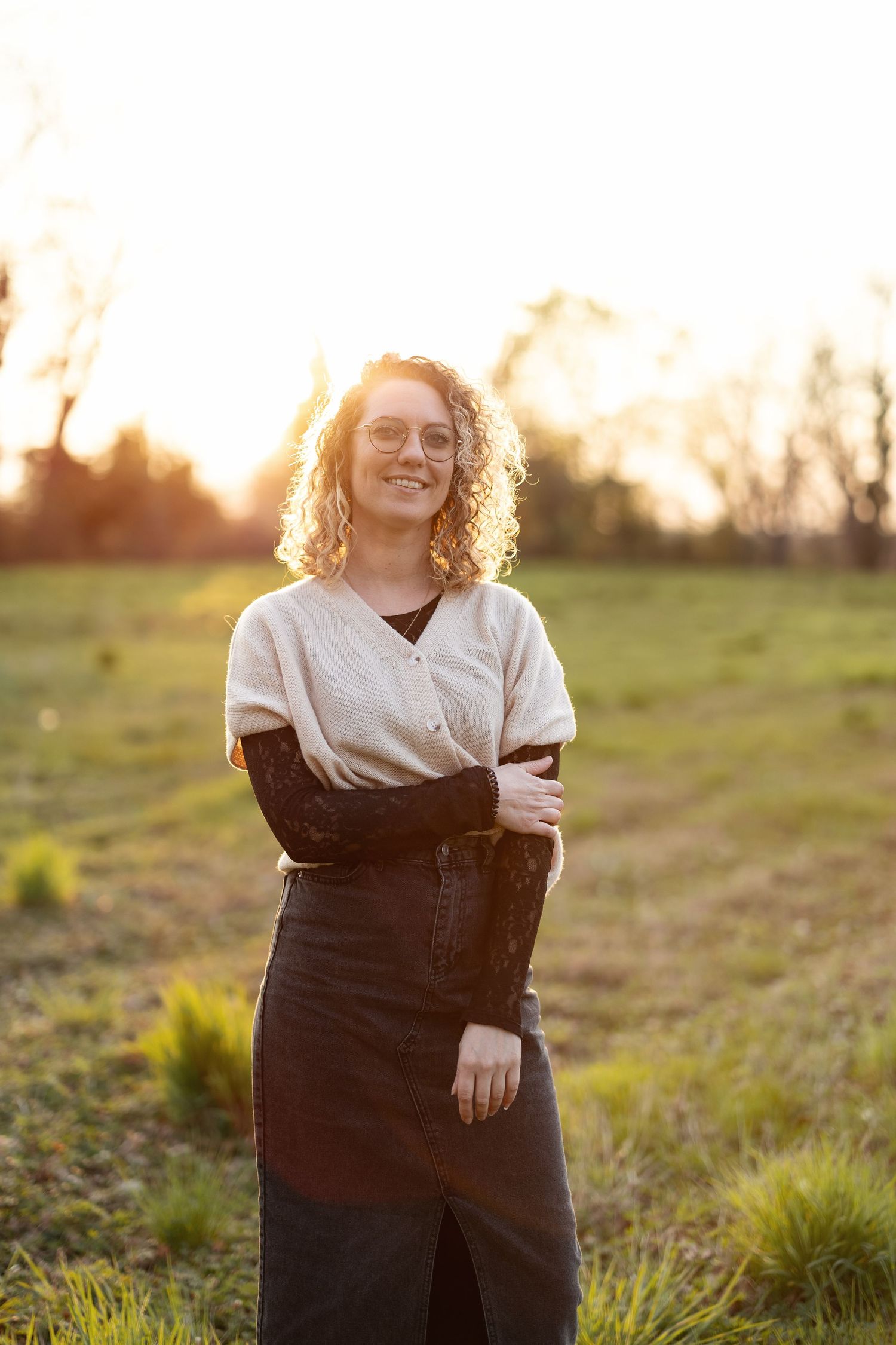 Smiling woman with curly hair and glasses standing in a sunlit green field at golden hour.