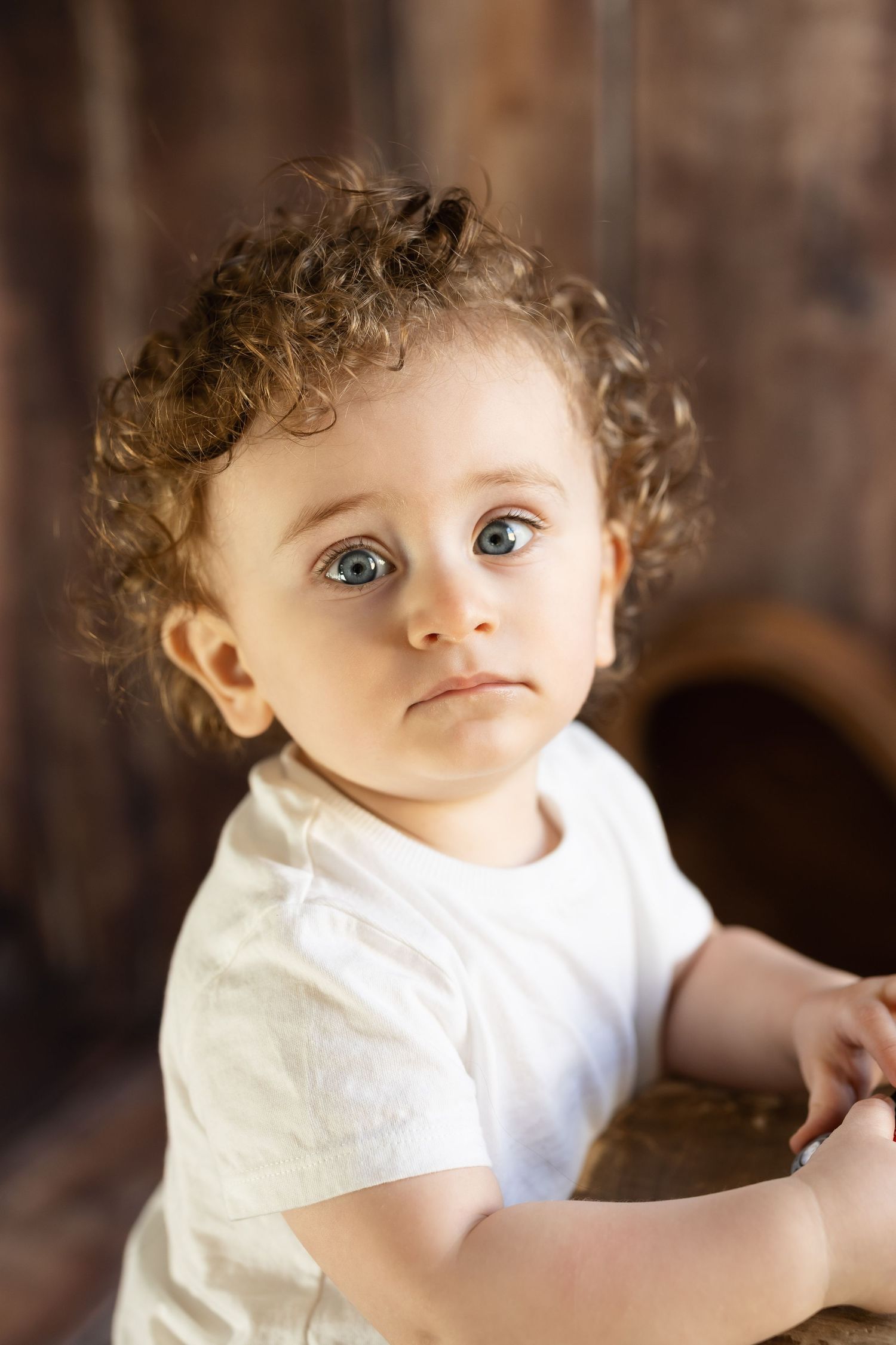 Adorable curly-haired baby with big blue eyes looking up in rustic wooden setting. photographe grossesse chavanoz lyon