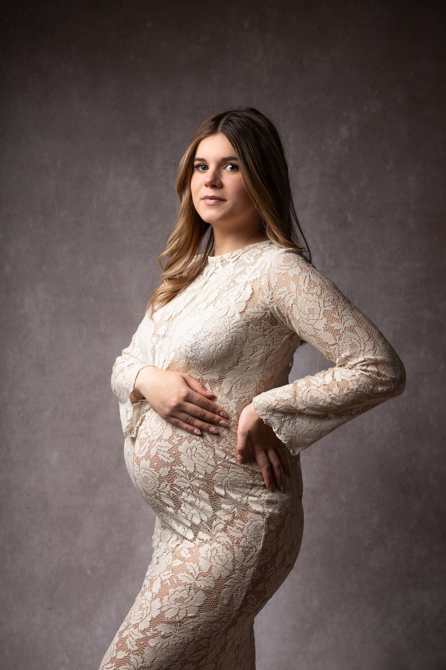 Elegant pregnant woman in white lace gown cradling baby bump, posing in studio against gray textured backdrop. photographe grossesse