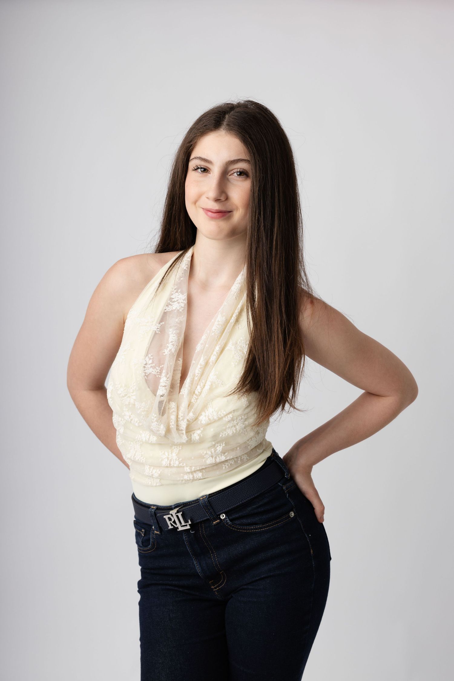 Young woman in cream halter top and dark jeans with Ralph Lauren belt, posing confidently against gray background.