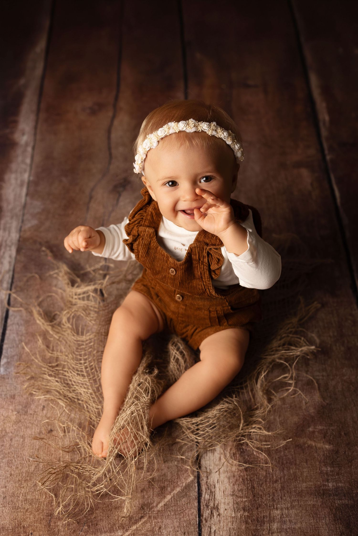Adorable baby girl in brown romper and floral headband sitting on rustic wood floor, smiling and reaching out.