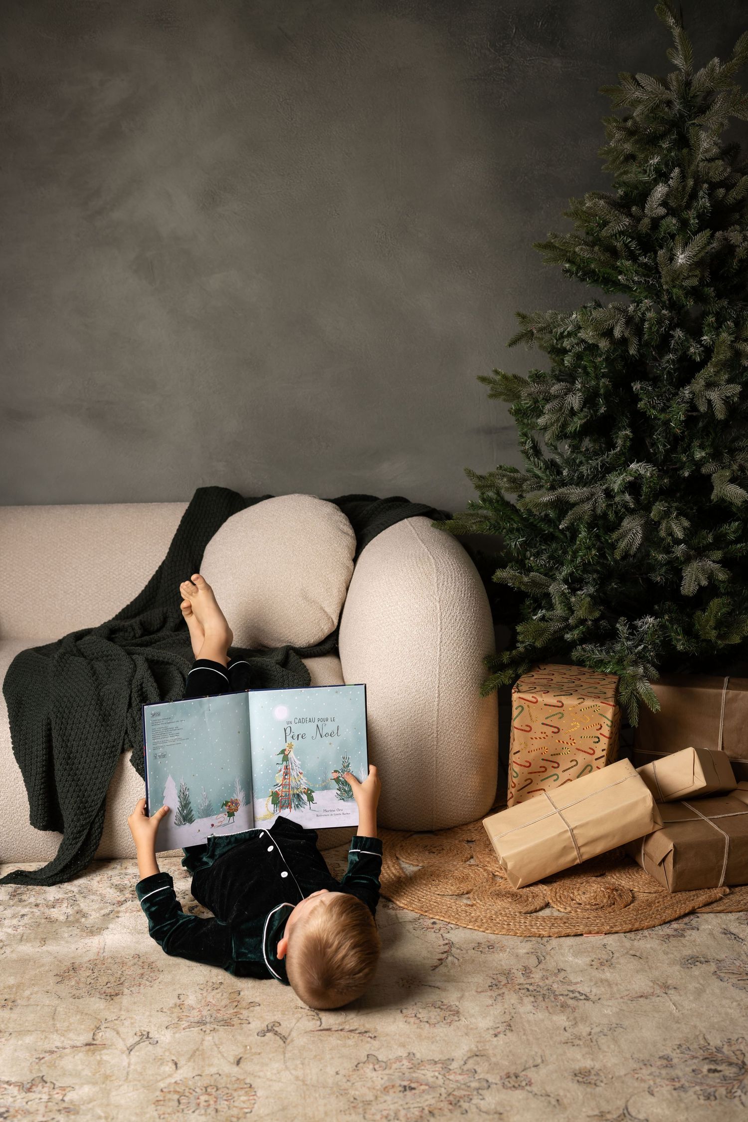 Child reading a Christmas book on the floor near a decorated tree and wrapped gifts in a cozy holiday setting.