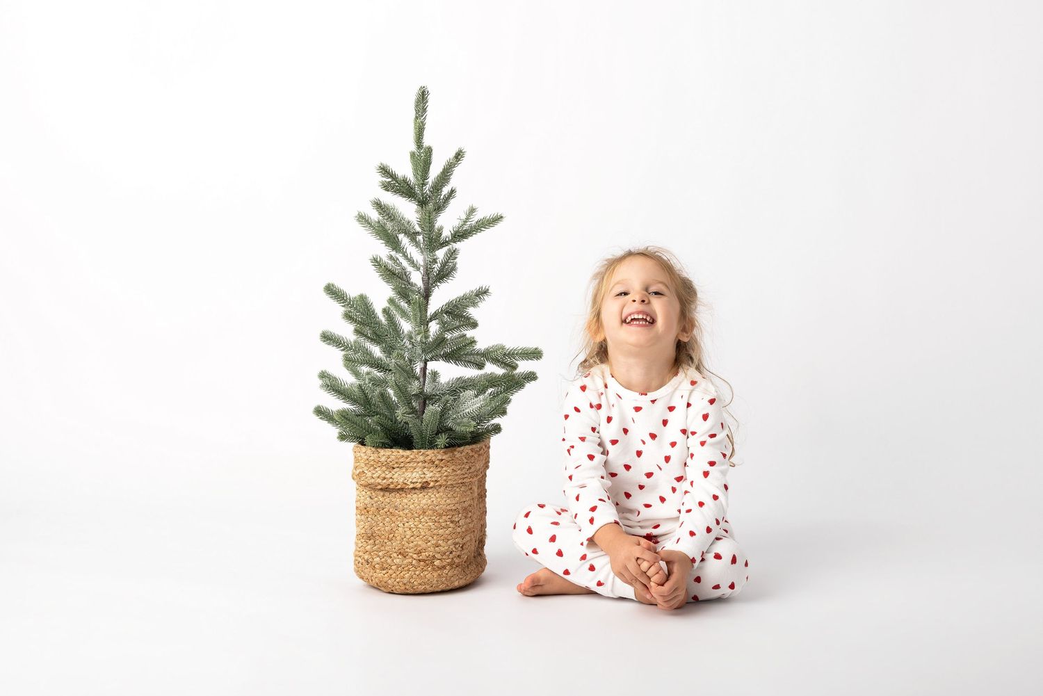Happy toddler girl in heart pajamas laughing beside a small potted Christmas tree on white background.
