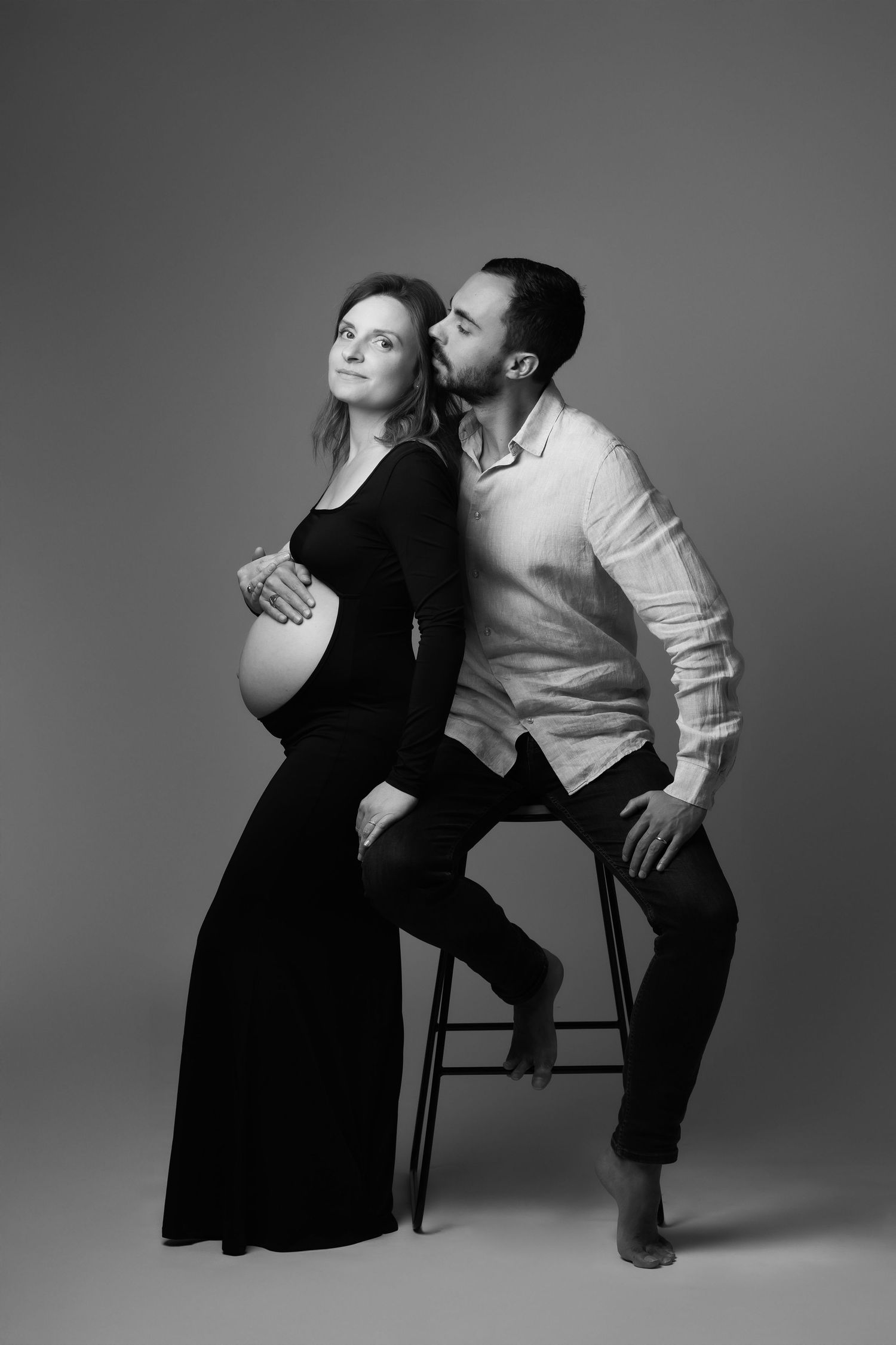 Black and white photo of a man kissing pregnant woman in black dress while sitting on a stool.