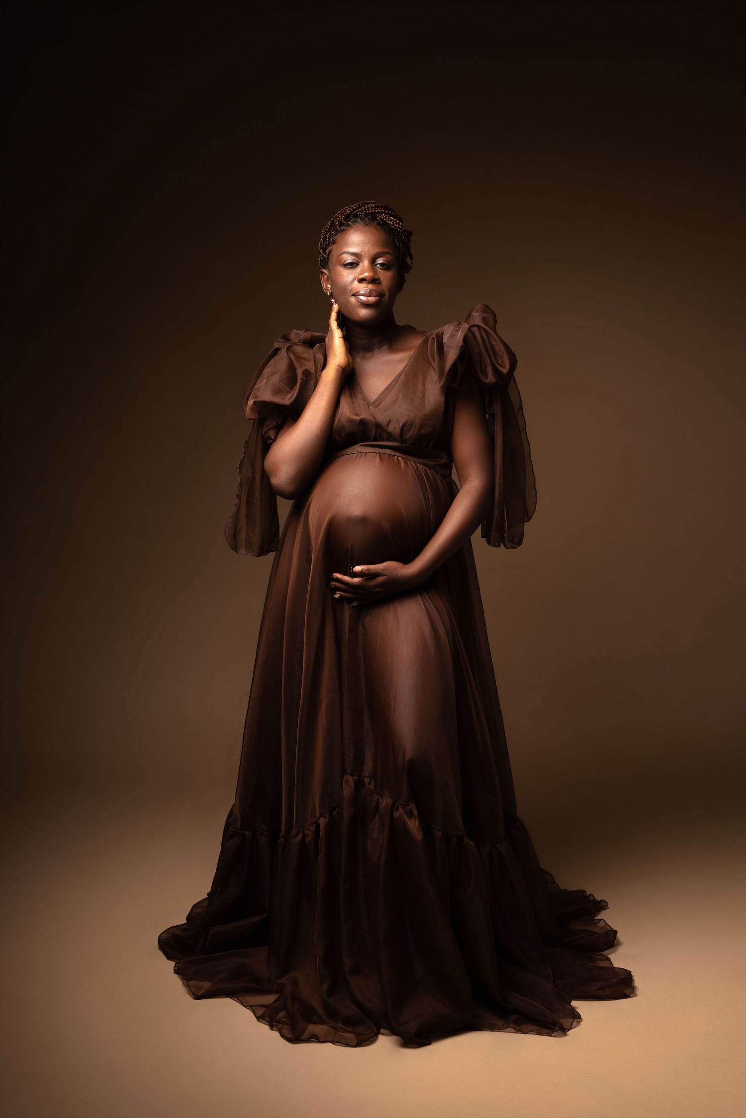 Pregnant woman in sheer brown maternity gown poses gracefully against warm brown studio background.