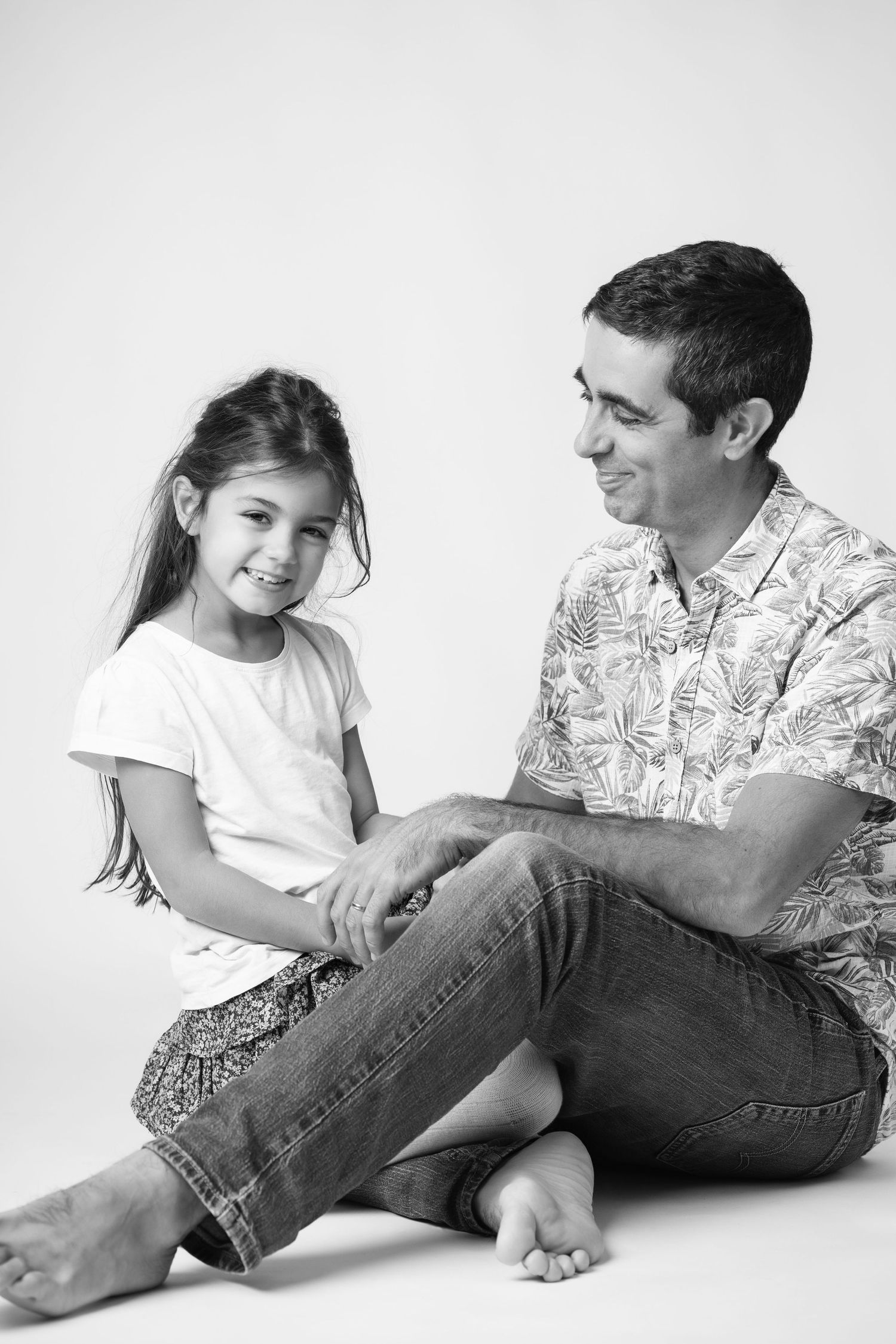 A man and young girl sit together on the floor, smiling in a black and white studio portrait.