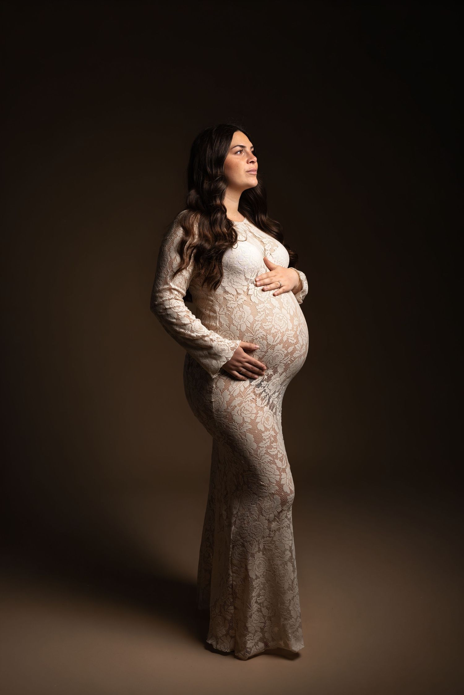 Pregnant woman in white lace gown cradling baby bump, studio portrait with dark background.