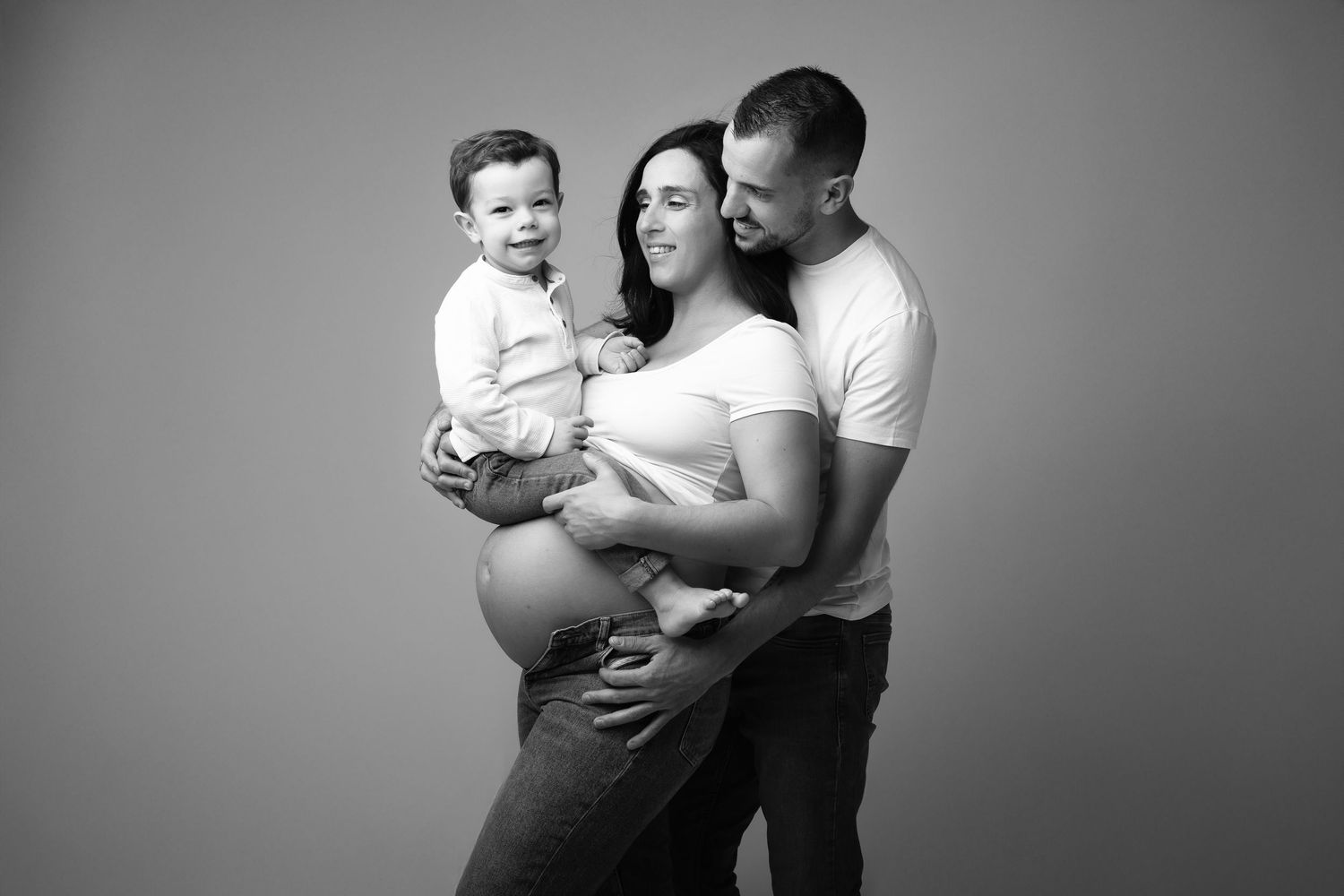 Black and white maternity photo of a pregnant woman with her partner and toddler son in a studio setting.
