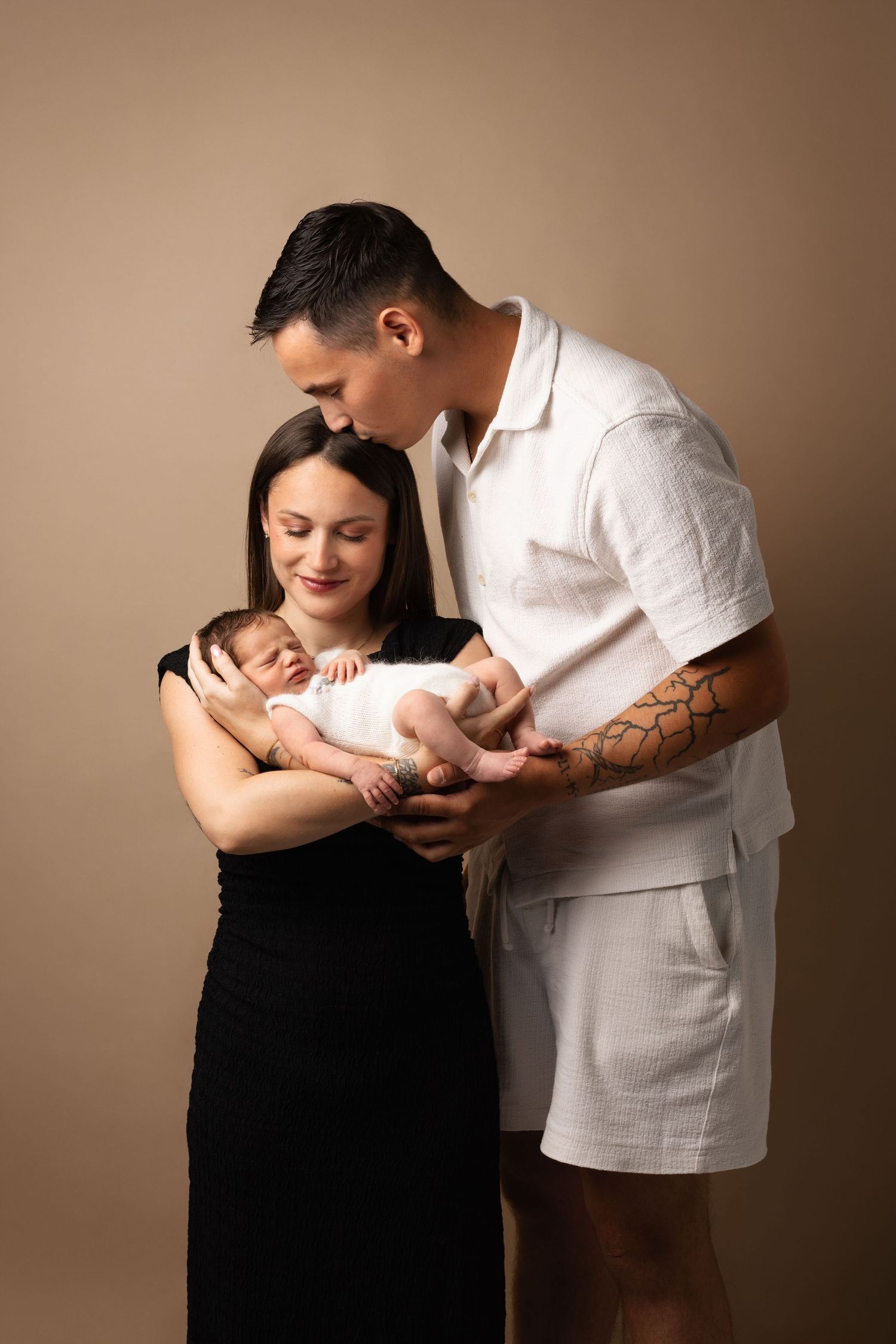A couple in a photography studio tenderly holds their newborn baby, the man kissing the woman's head.
