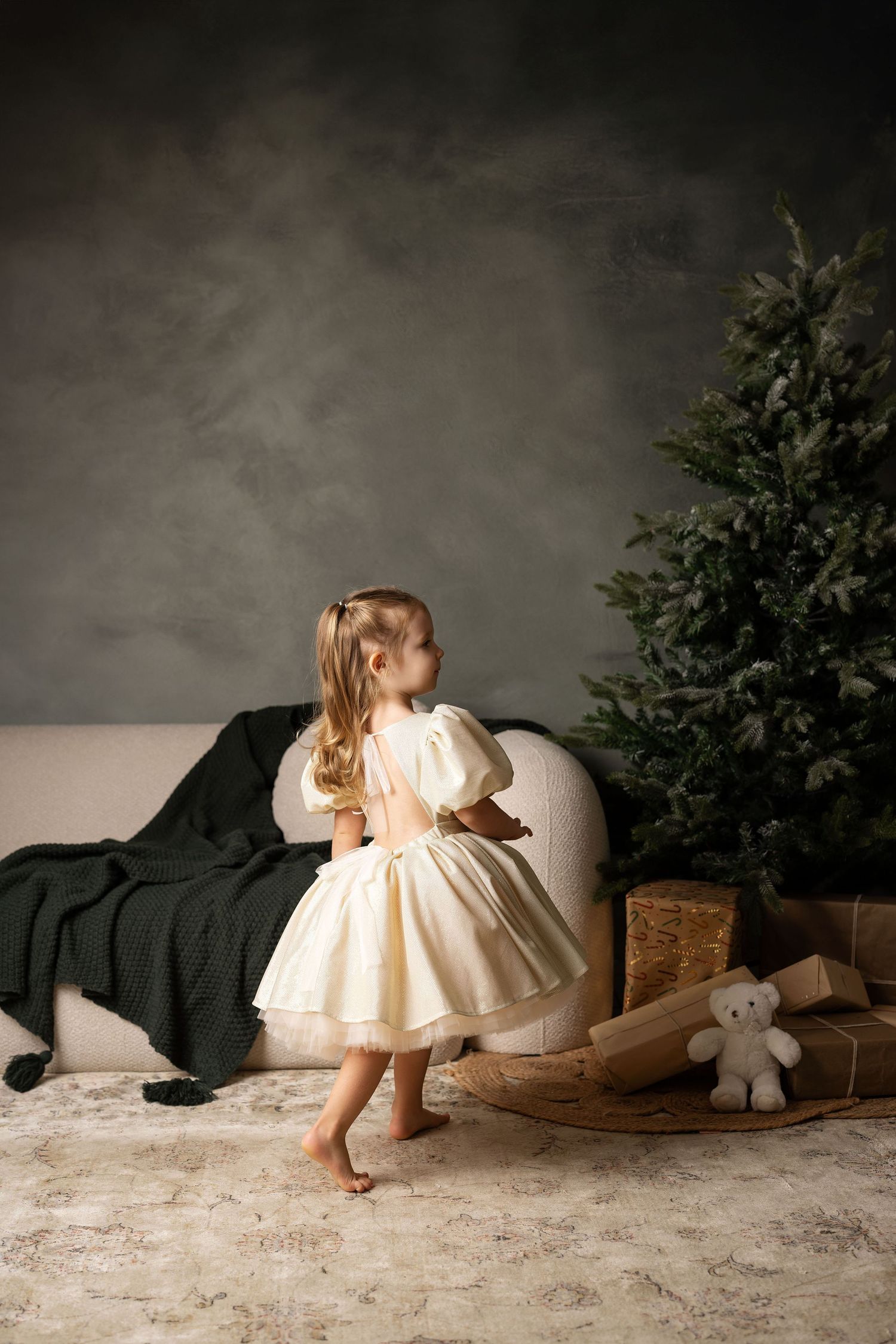 Young girl in cream dress stands barefoot by Christmas tree with gifts and teddy bear in festive room.