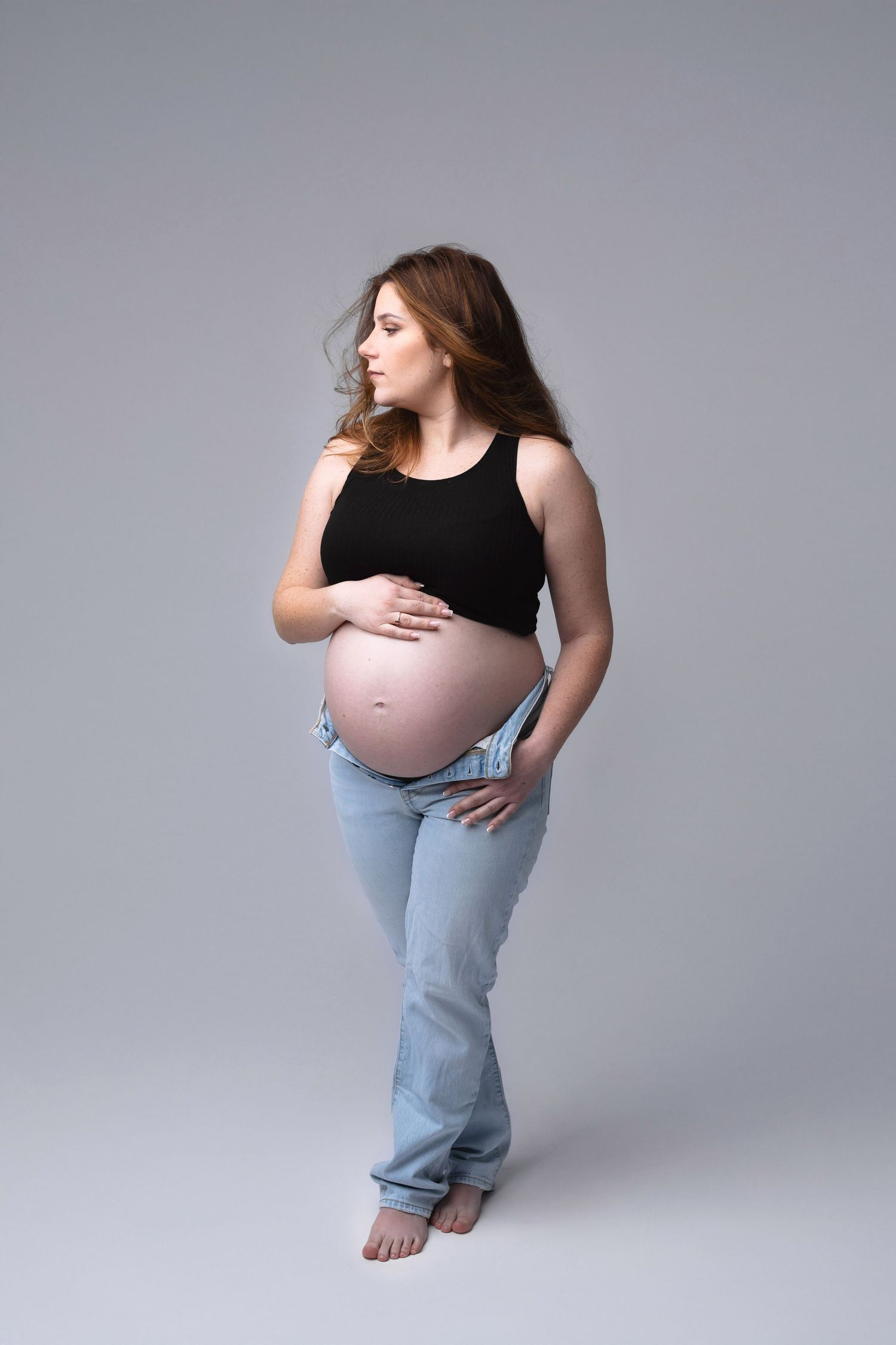 Pregnant woman in black tank top and unzipped jeans showing bare baby bump in studio. photographe grossesse chavanoz lyon