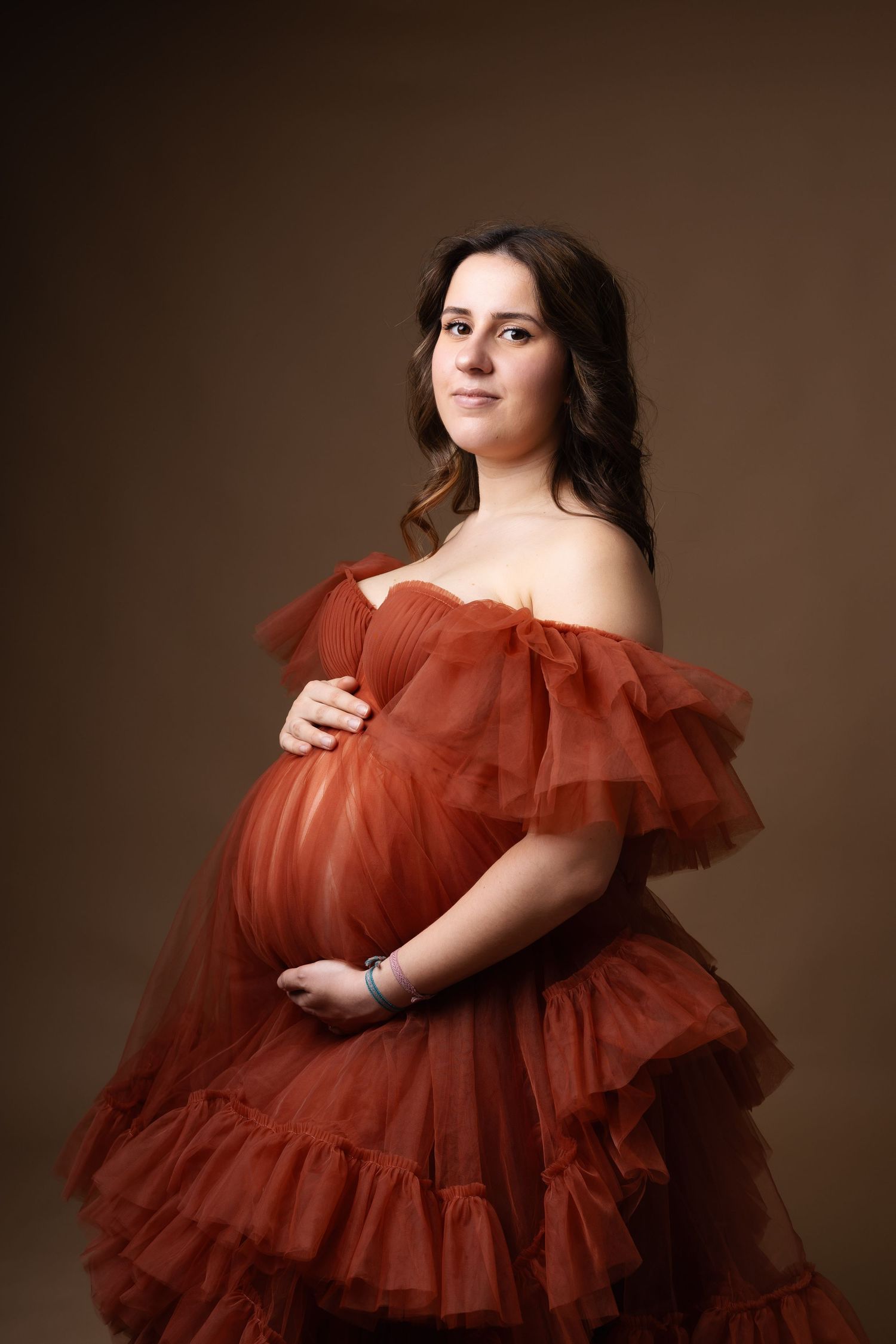 Beautiful pregnant woman in rust orange ruffled tulle gown holding baby bump on brown background. photographe grossesse chavanoz lyon