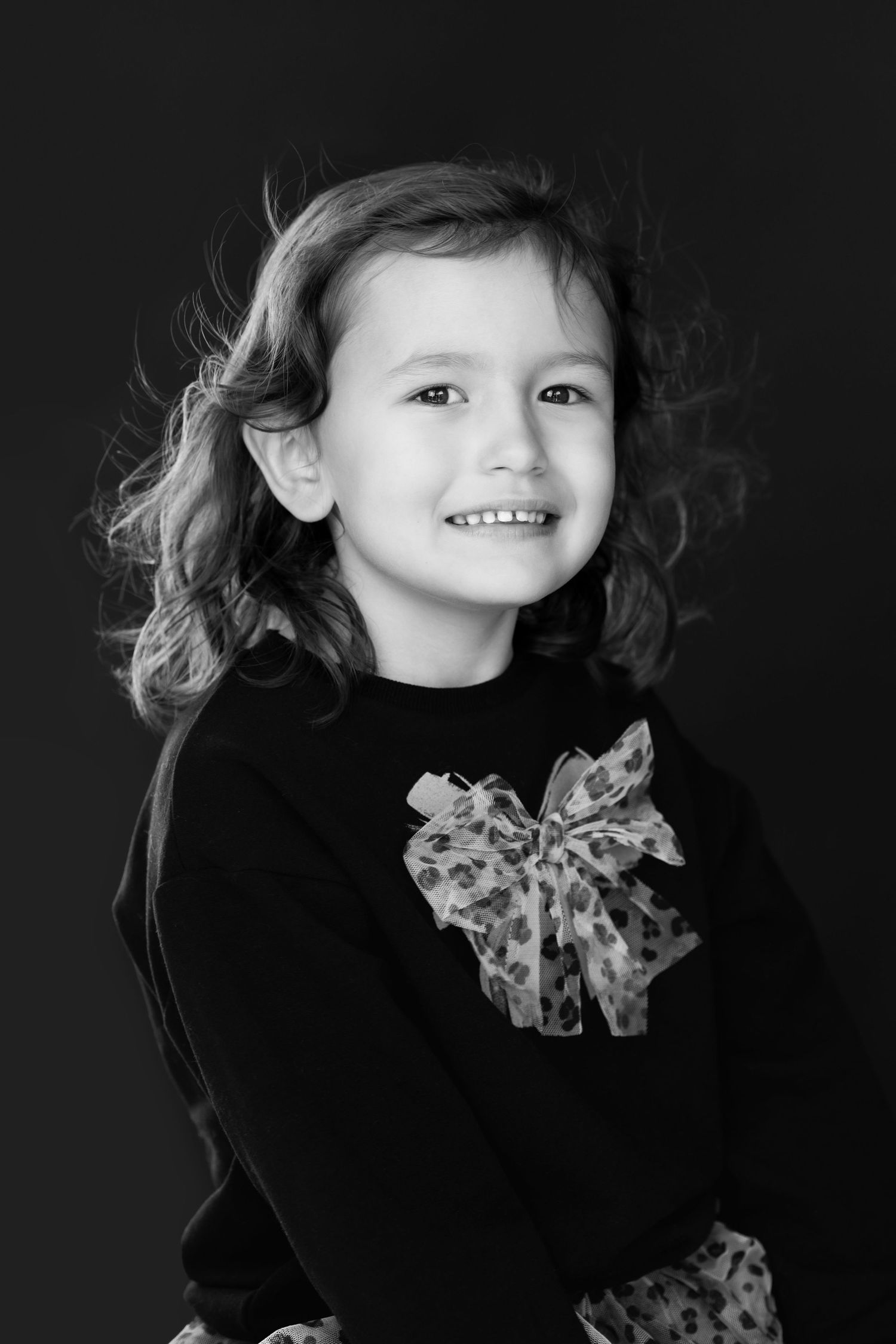 Adorable smiling little girl with curly hair wearing black top with bow in black and white portrait.