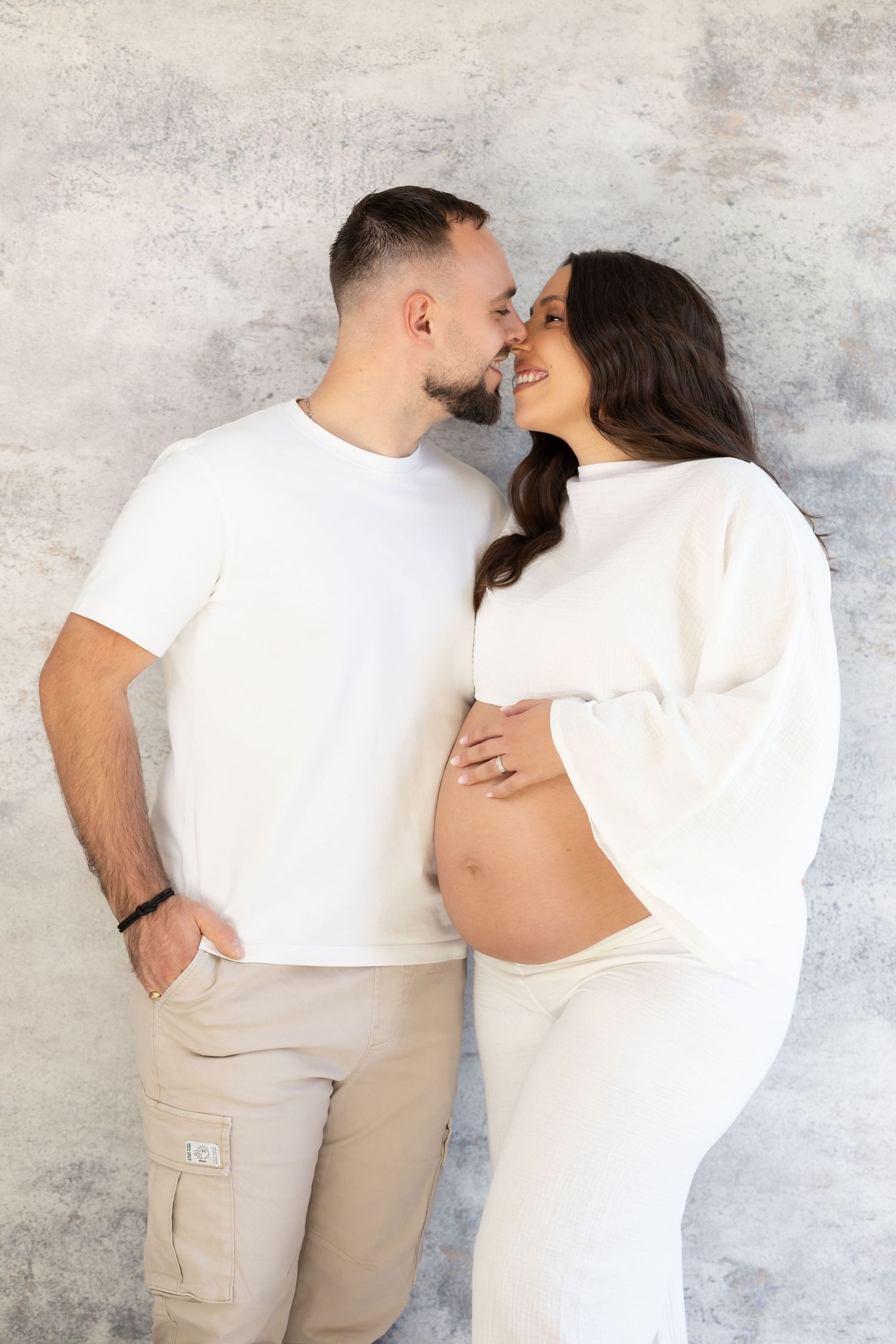 Couple sharing a tender moment, pregnant woman in white outfit showing bare bump against grey wall.